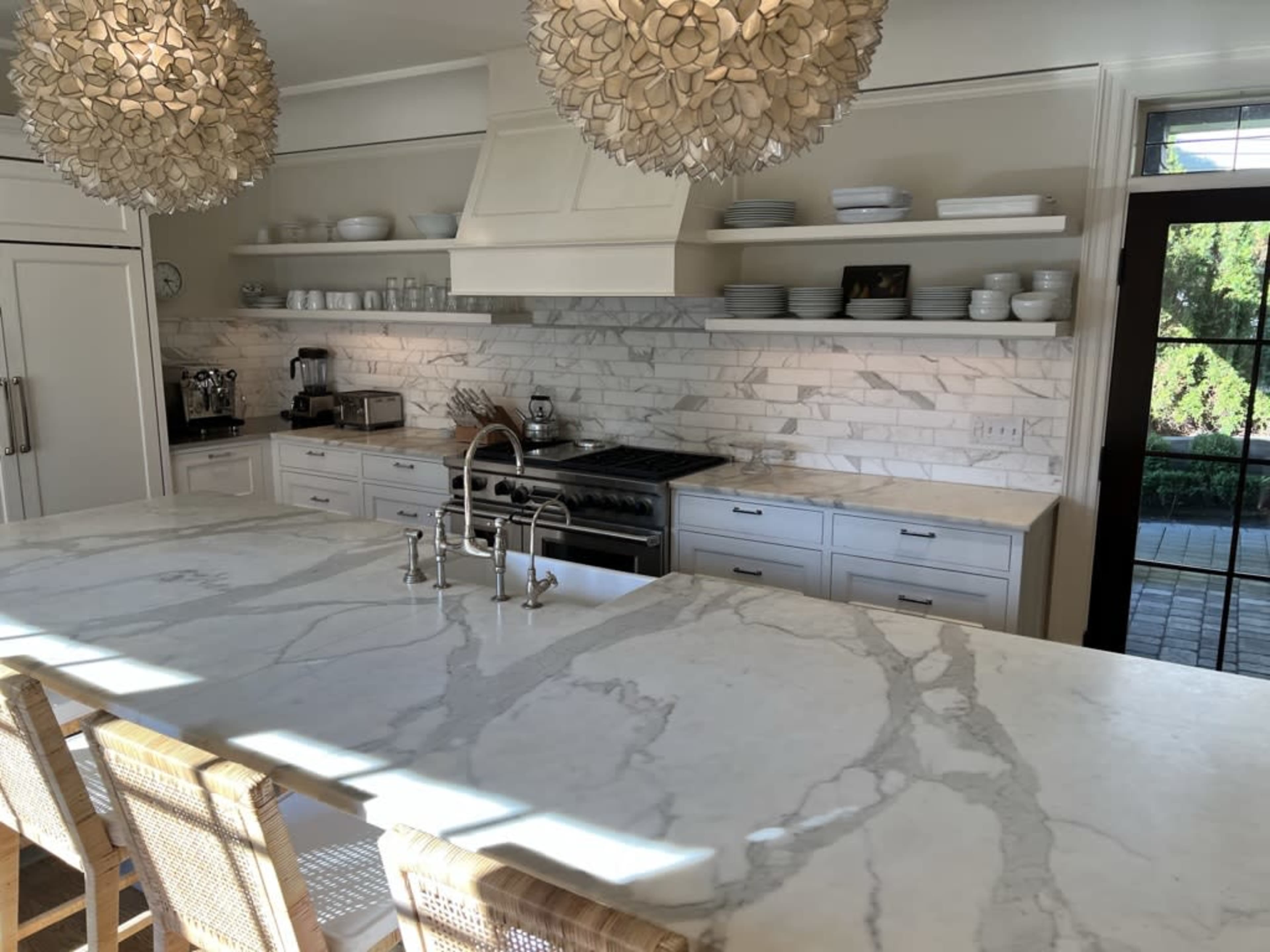 A modern kitchen features white cabinetry, a large marble island with seating, and stone backsplash with kitchen appliances and dishware neatly arranged on shelves.