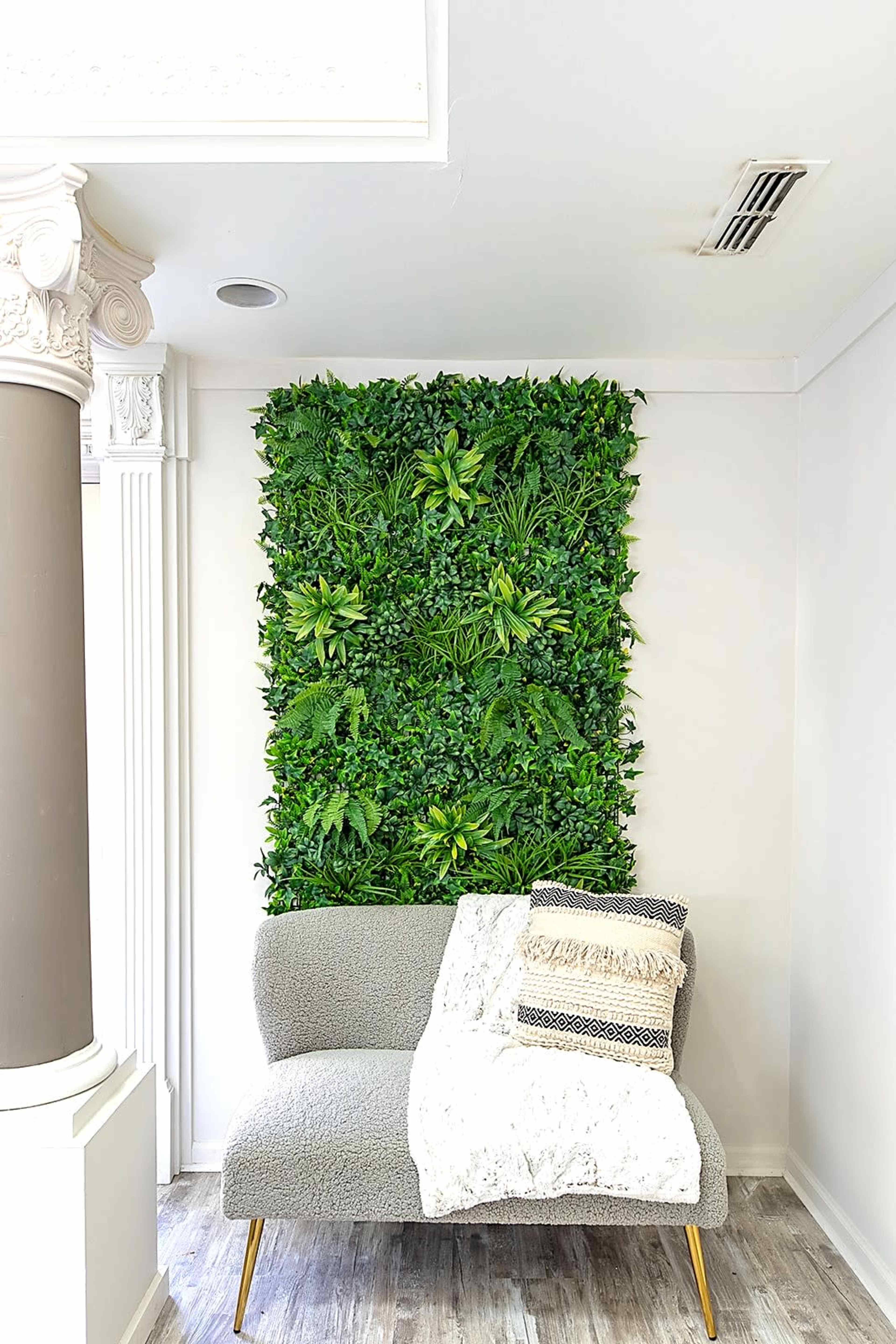 A gray sofa with a patterned throw pillow is placed against a green wall of artificial plants in a well-lit interior space.