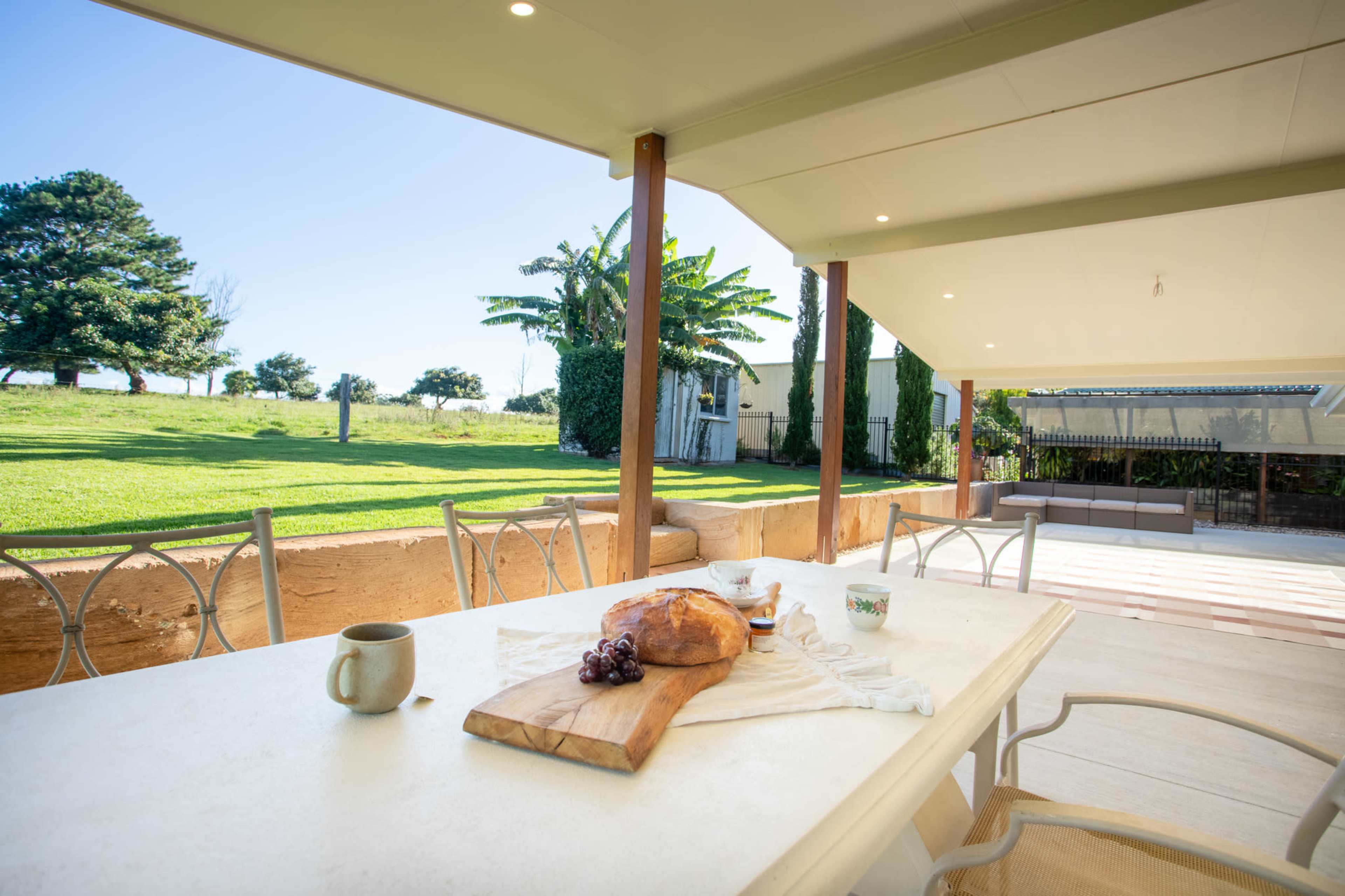 A table is set on a patio with a loaf of bread, a cup, and grapes, overlooking a grassy yard with trees in the background.