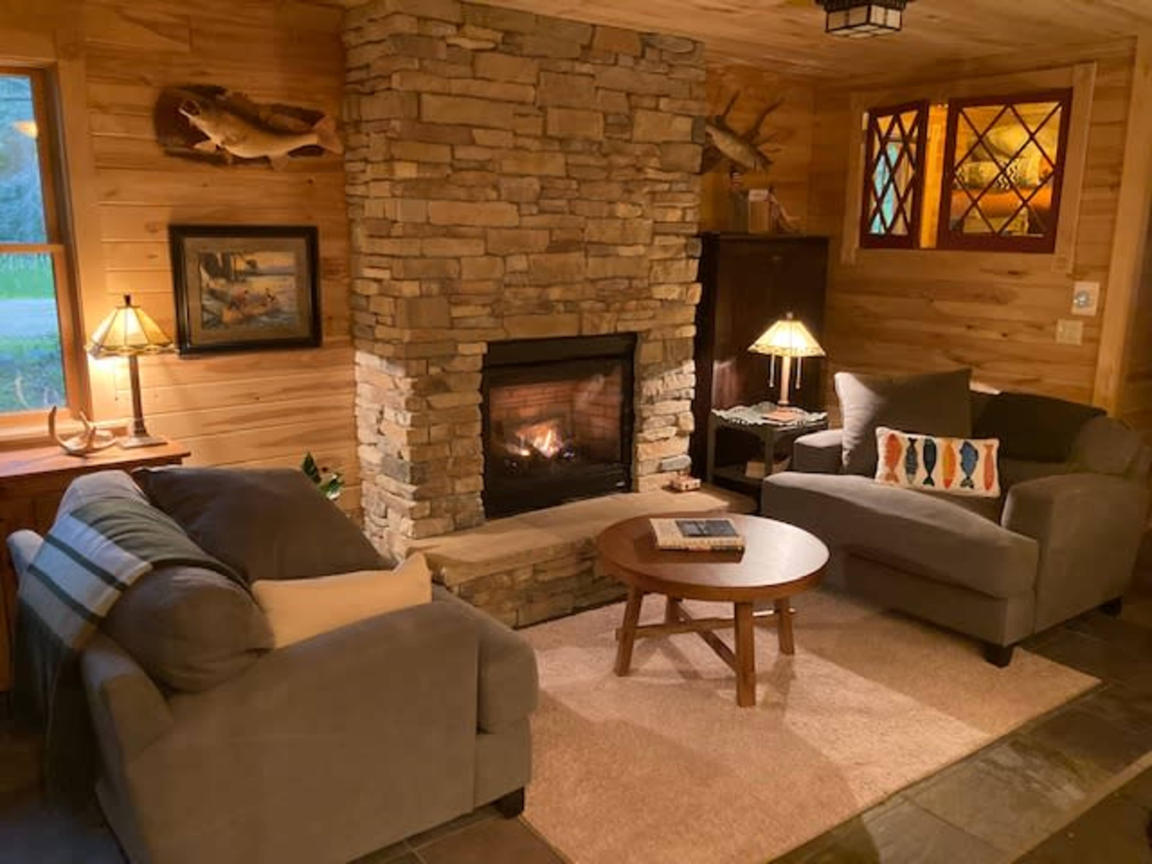 The image shows a cozy living room with a stone fireplace, two armchairs, a coffee table, and wooden decor.