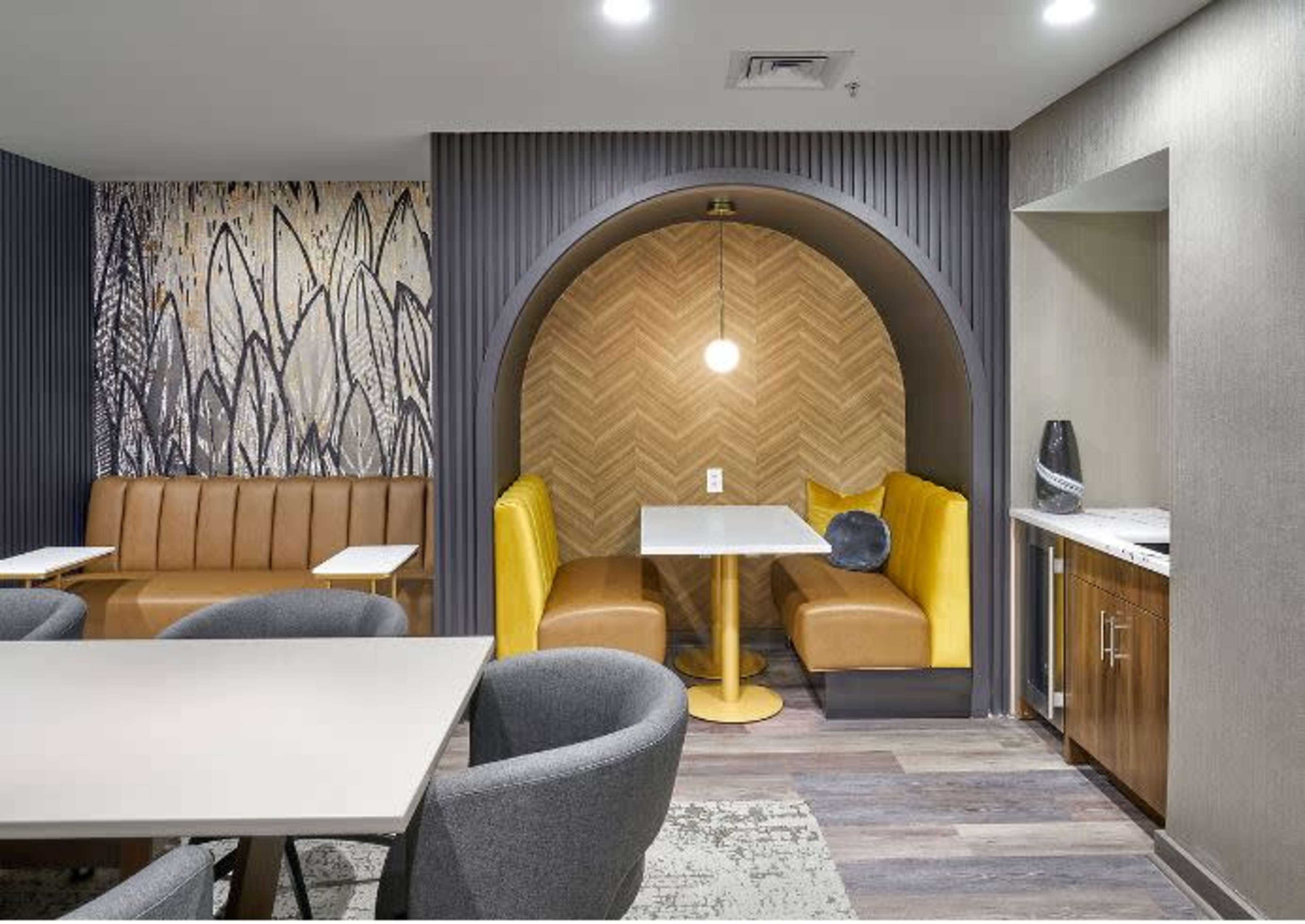 The image shows a modern lounge area with a curved booth featuring yellow seating and a round table, adjacent to a textured wall and a kitchenette.