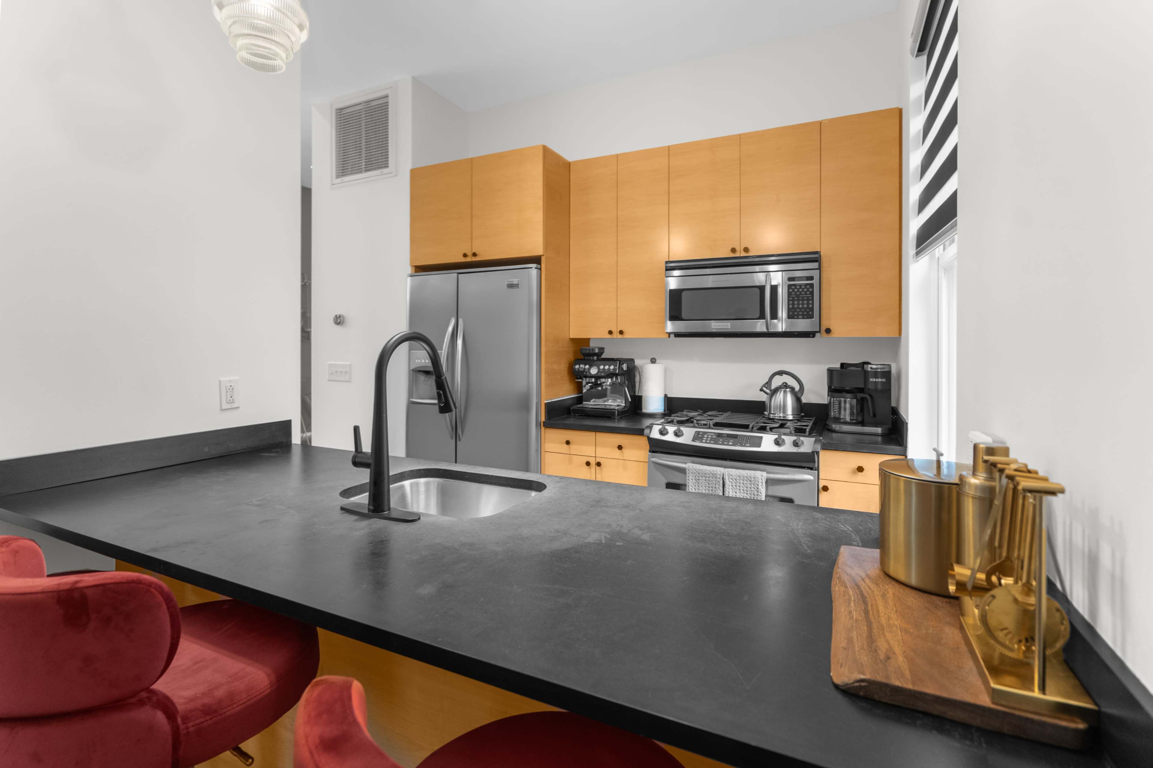 A modern kitchen features wooden cabinets, stainless steel appliances, and a black countertop with a sink and red bar stools.