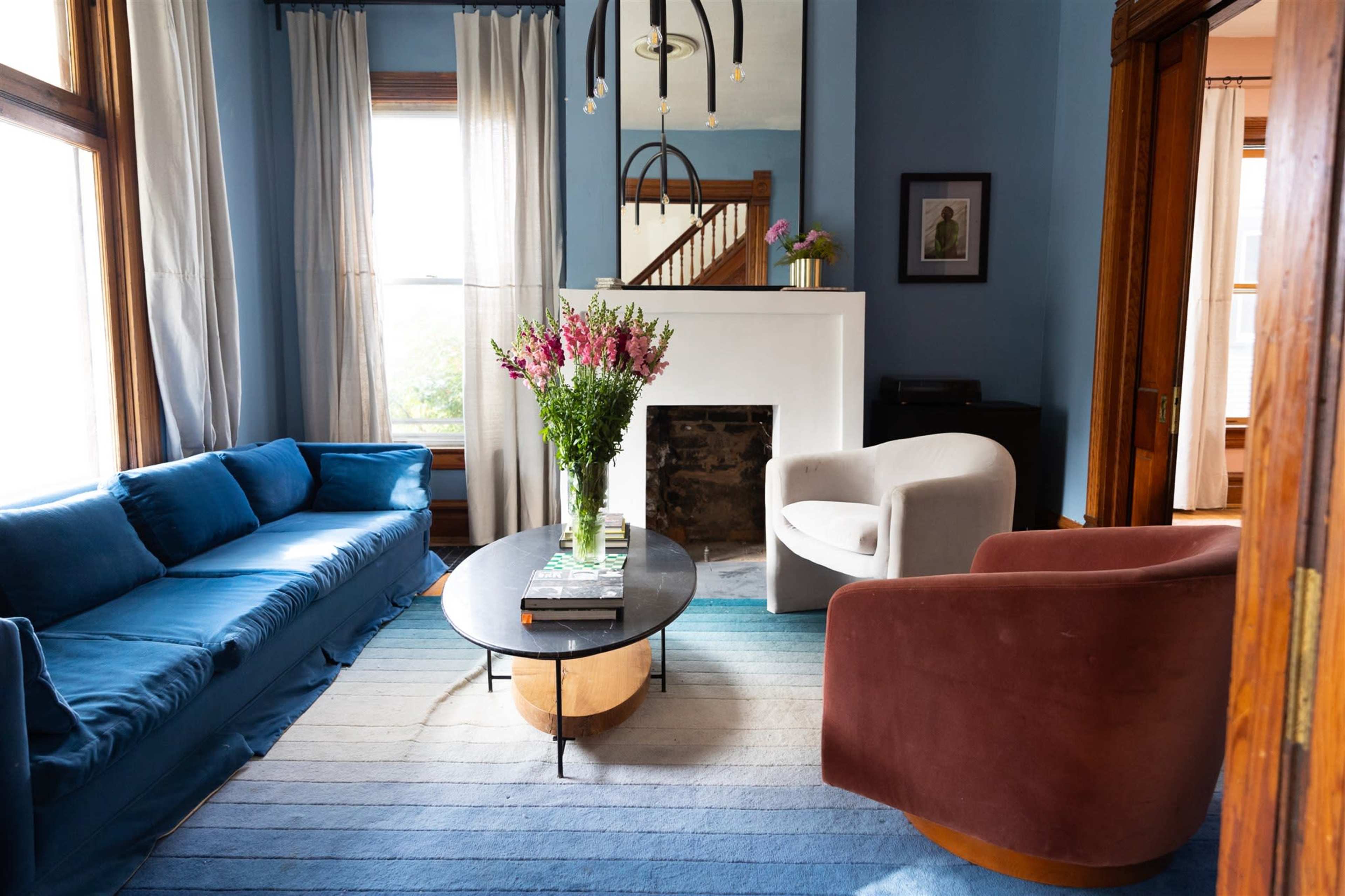 A living room with a blue couch, a white chair, a circular coffee table, and flowers in a vase on top of the table, with large windows providing natural light.