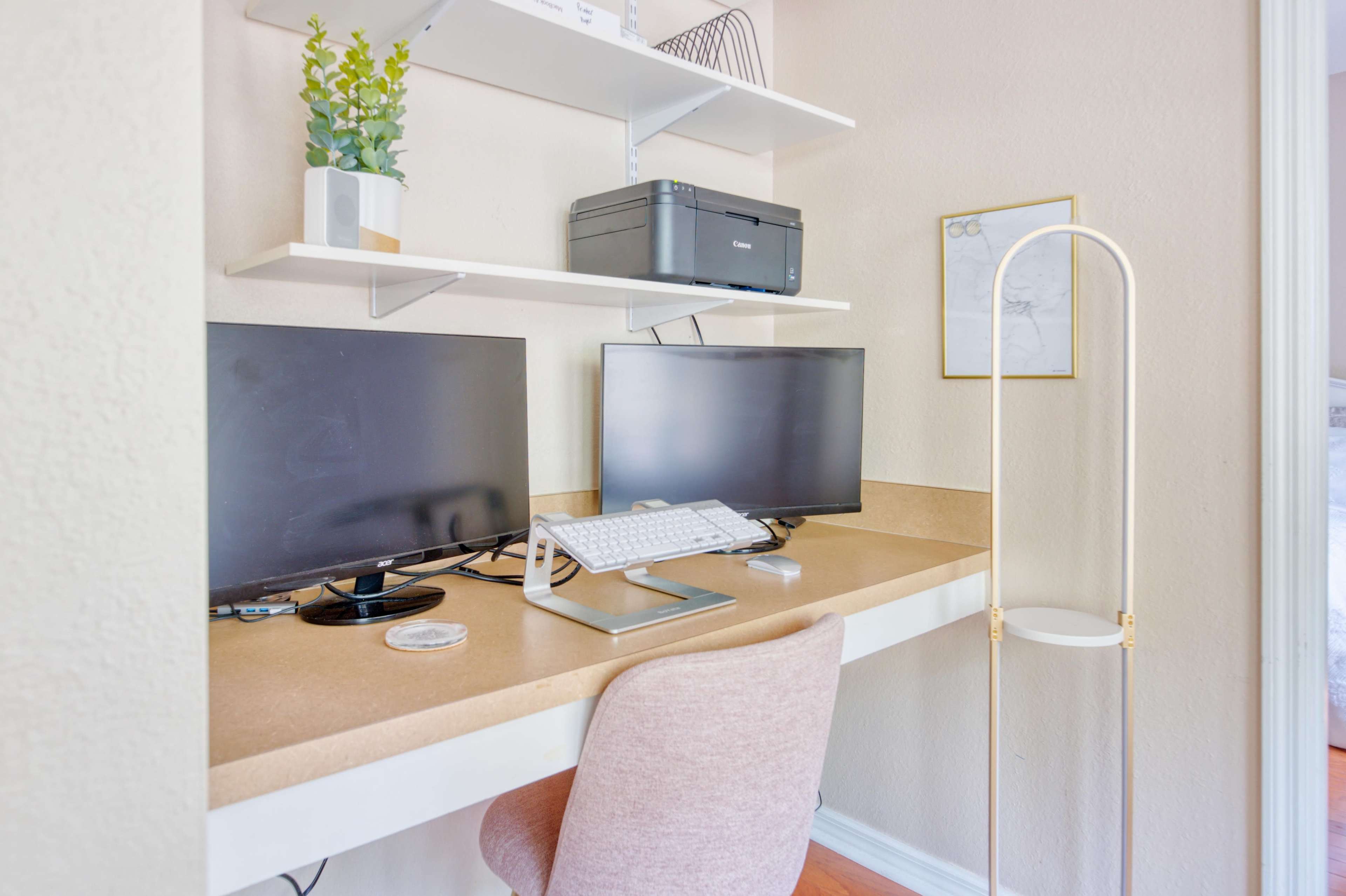 A workspace featuring two computer monitors, a keyboard on a stand, and a pink chair against a light-colored wall with shelves above holding decorative plants and a printer.