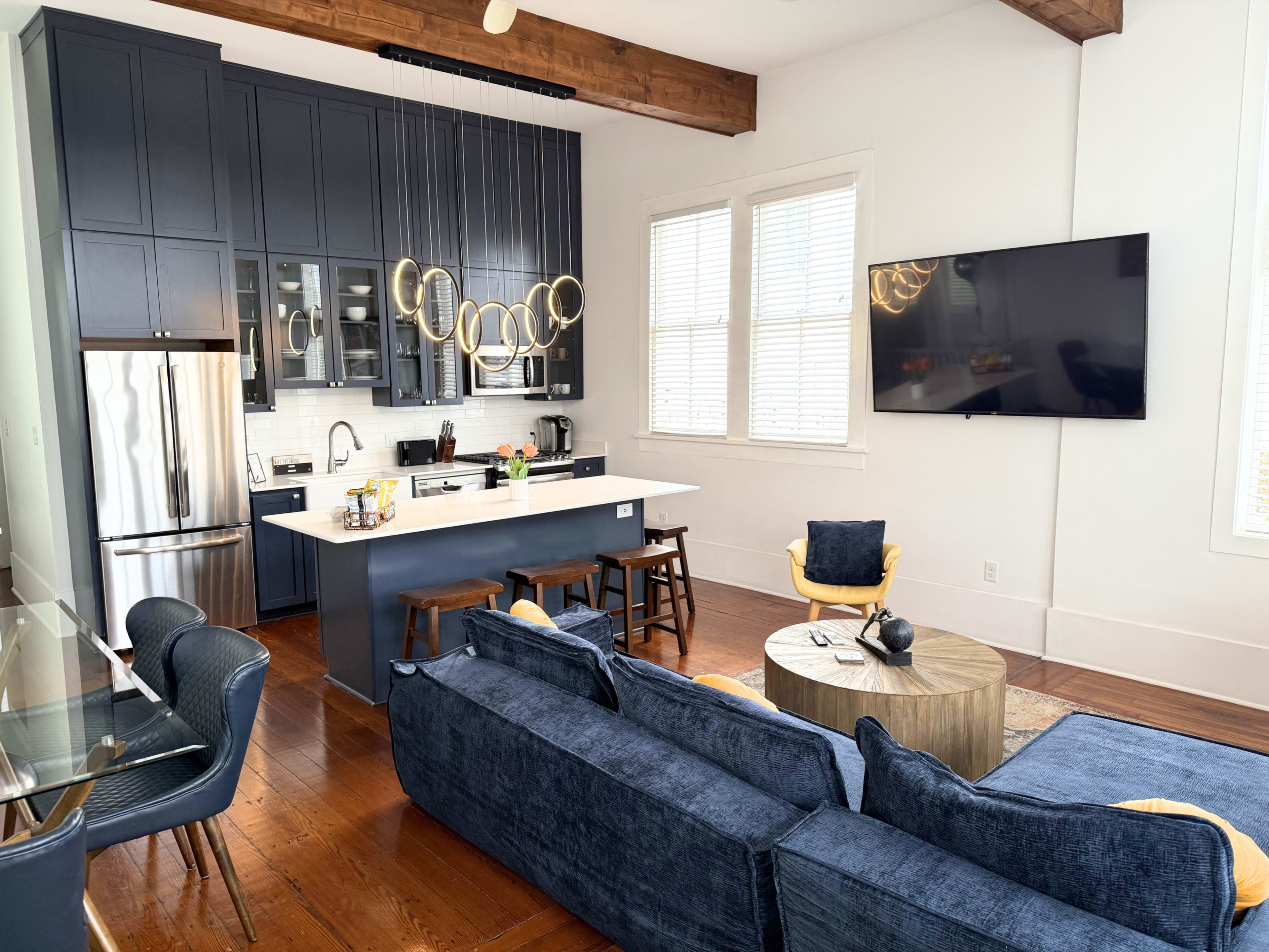 The image shows a modern kitchen and living area with dark cabinetry, stainless steel appliances, and a blue sofa arranged around a wooden coffee table.