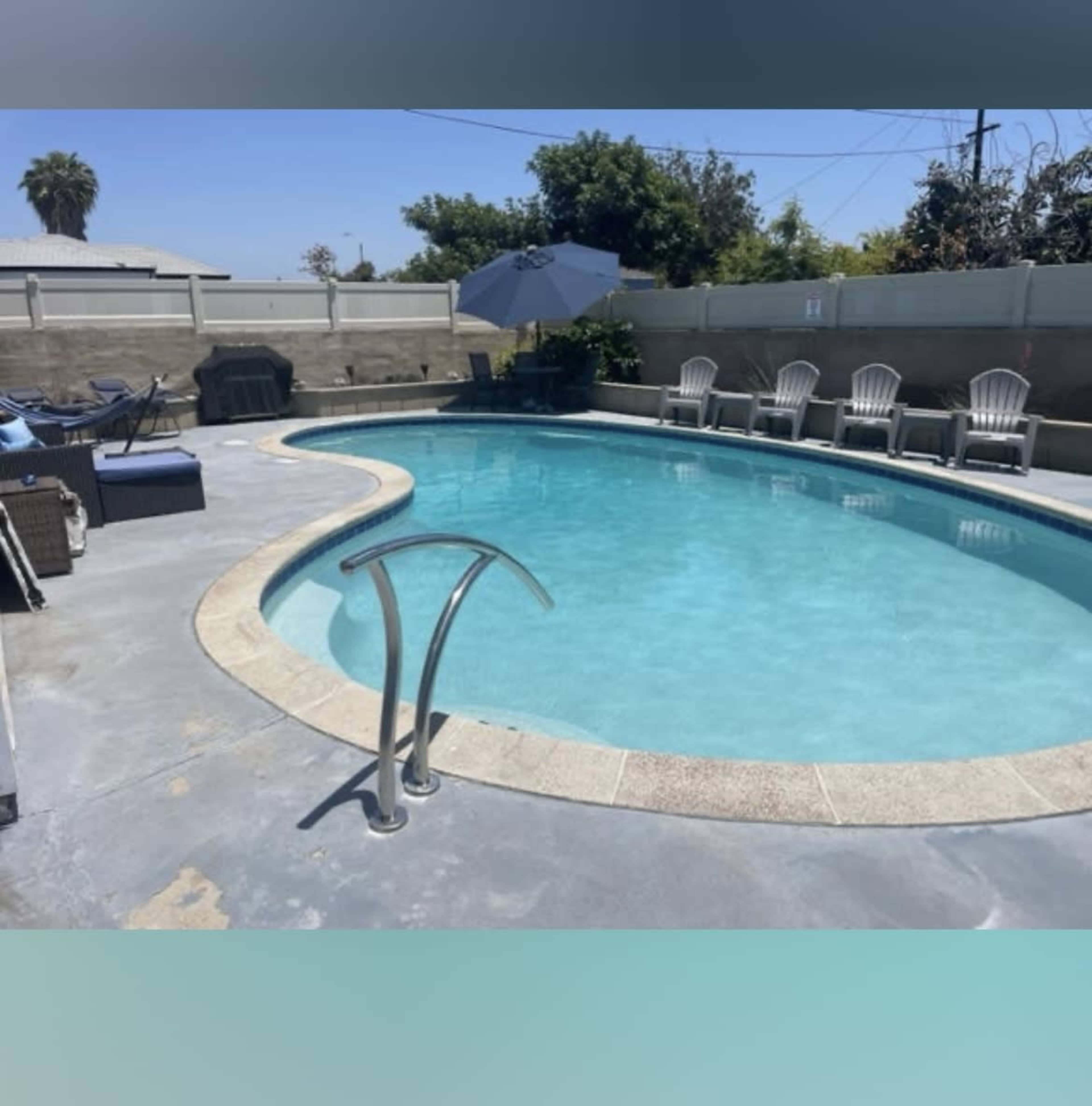 A rectangular swimming pool surrounded by patio chairs and an umbrella in a sunny outdoor area.