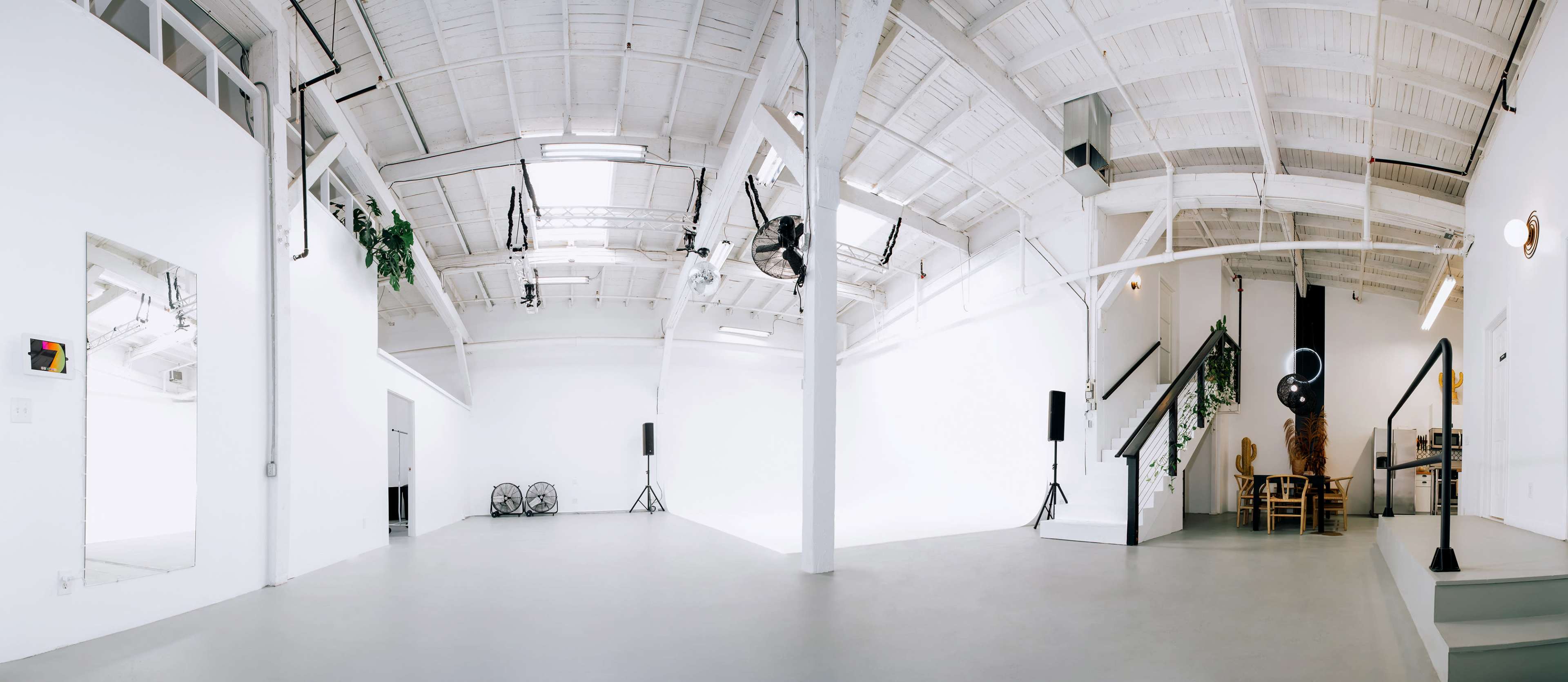 A spacious, well-lit studio with a high ceiling, white walls, and minimalistic decor, featuring speakers, lighting equipment, and a staircase leading to a small seated area.
