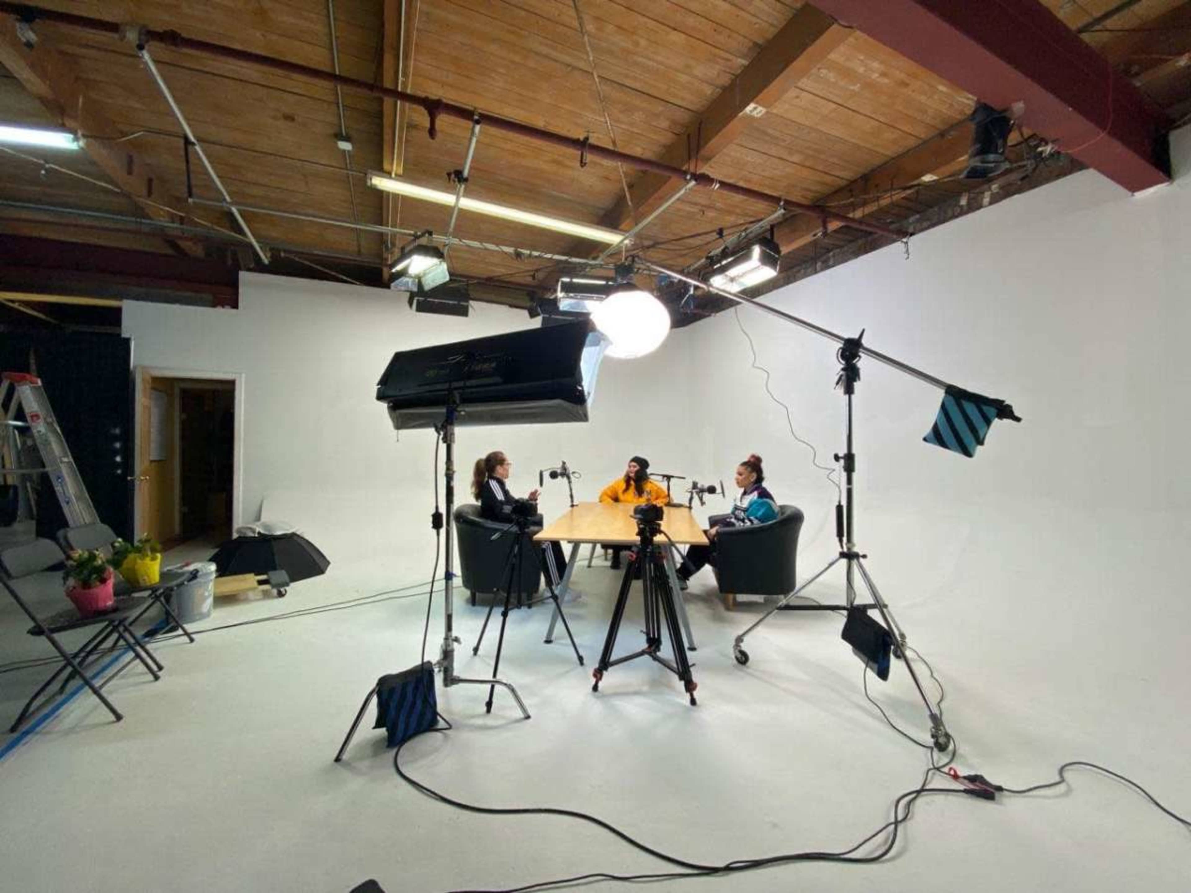 Two people sit at a table in a studio setting, surrounded by lighting equipment and cameras.