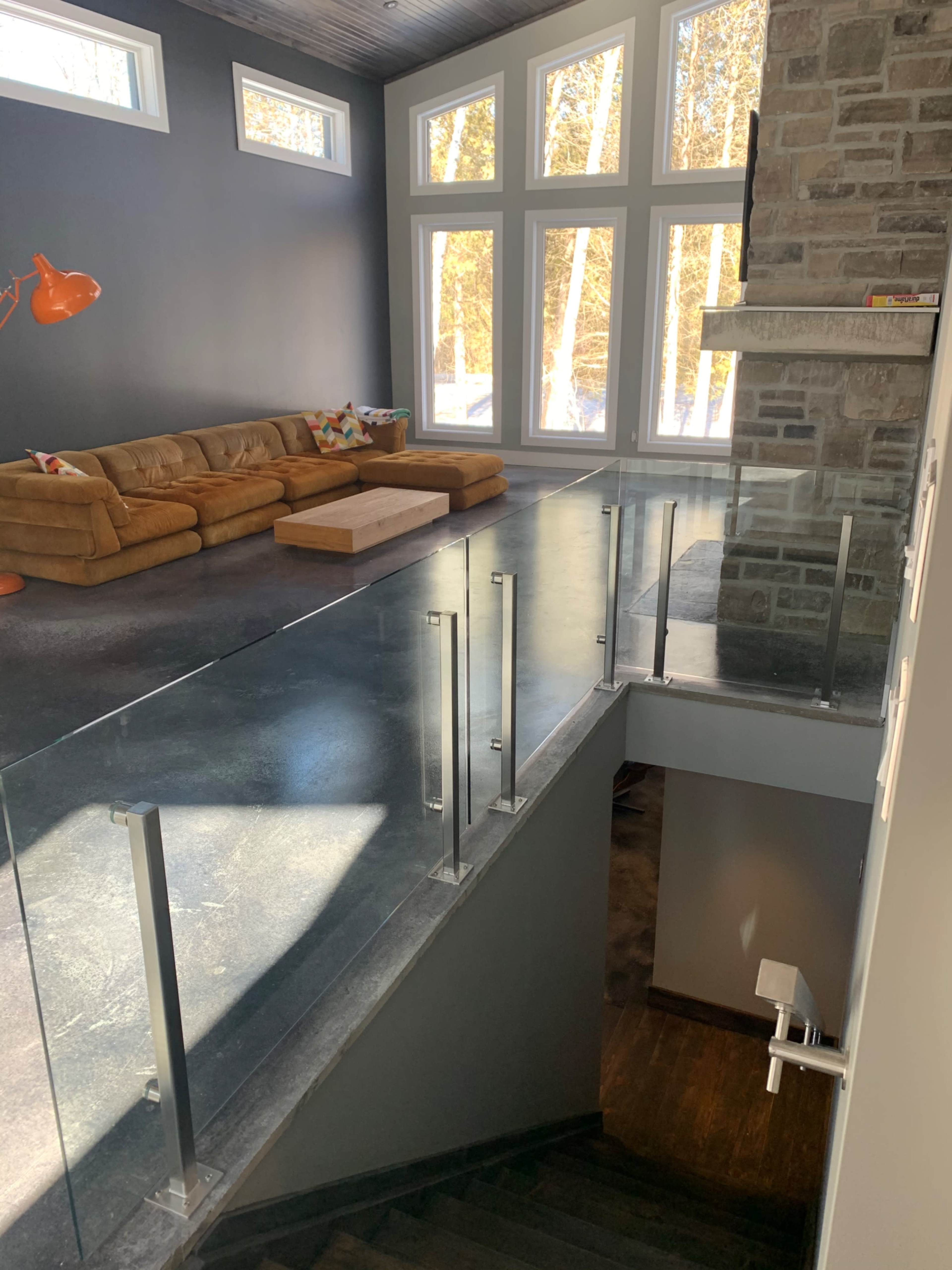 A modern living room with a large brown couch, a wooden coffee table, and a staircase leading down, all illuminated by tall windows allowing natural light to enter.