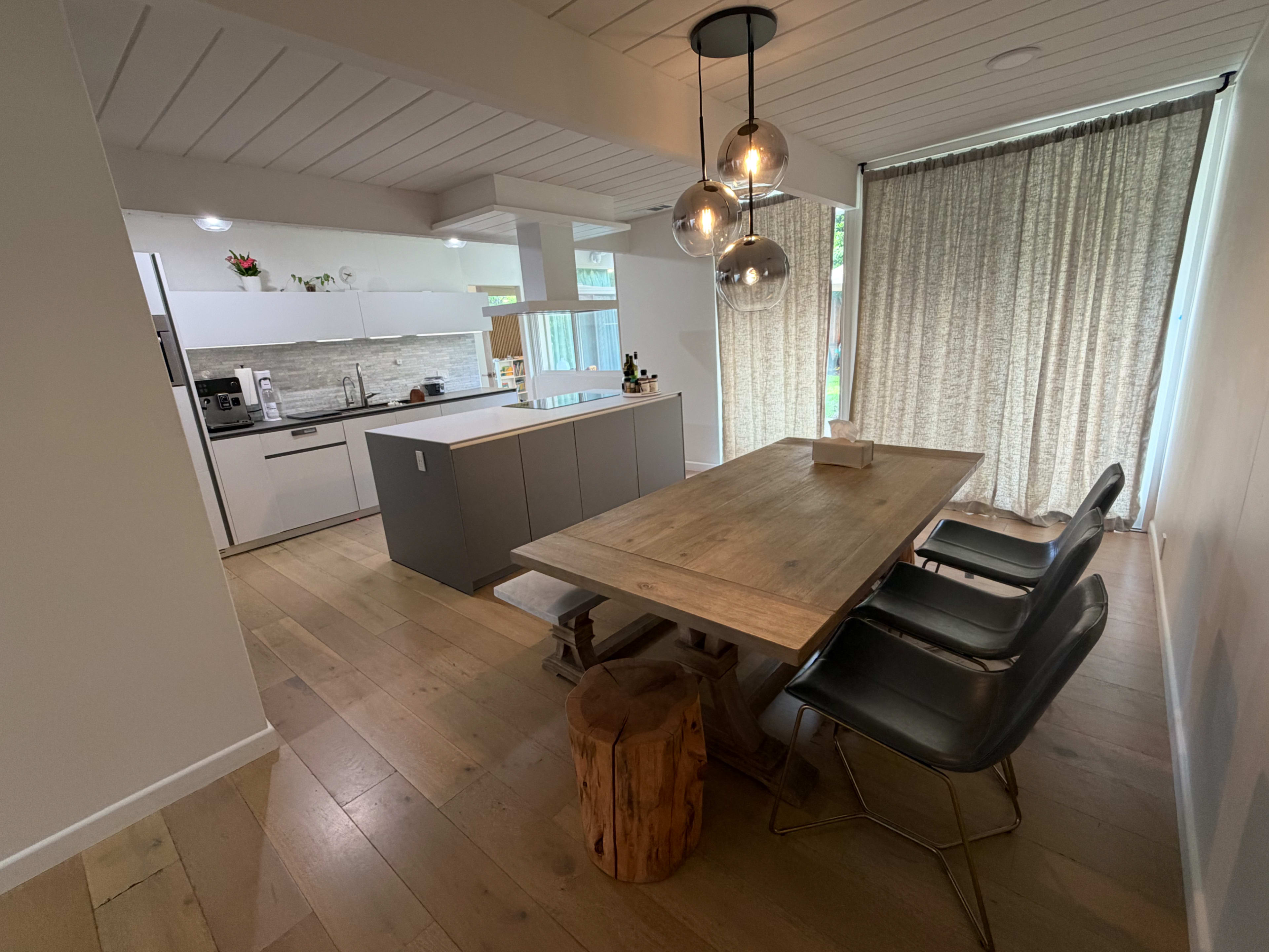 The image shows a modern kitchen and dining area featuring a wooden dining table, black chairs, a central kitchen island, and sheer curtains allowing natural light to enter.