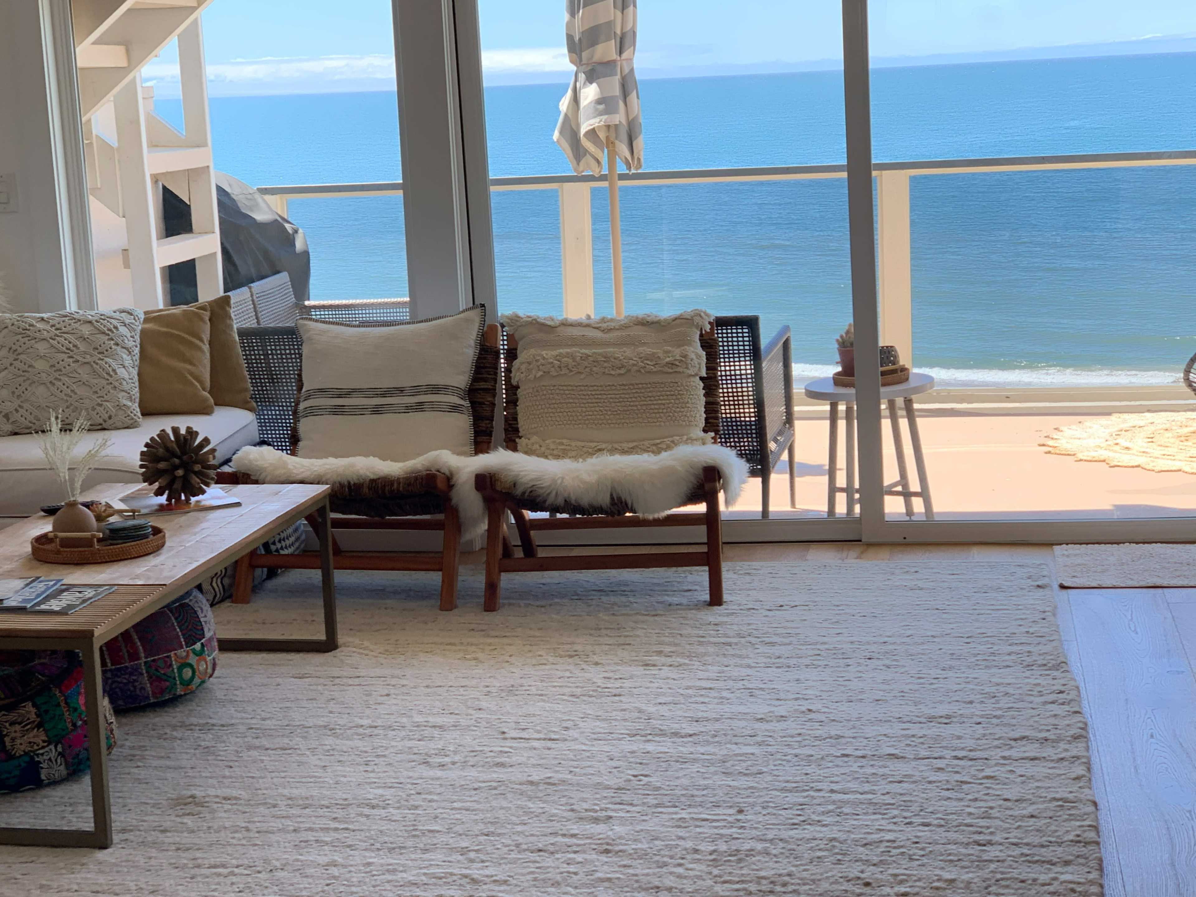 The scene features a cozy living area with two woven chairs, a wooden coffee table, and large windows overlooking a calm ocean view.