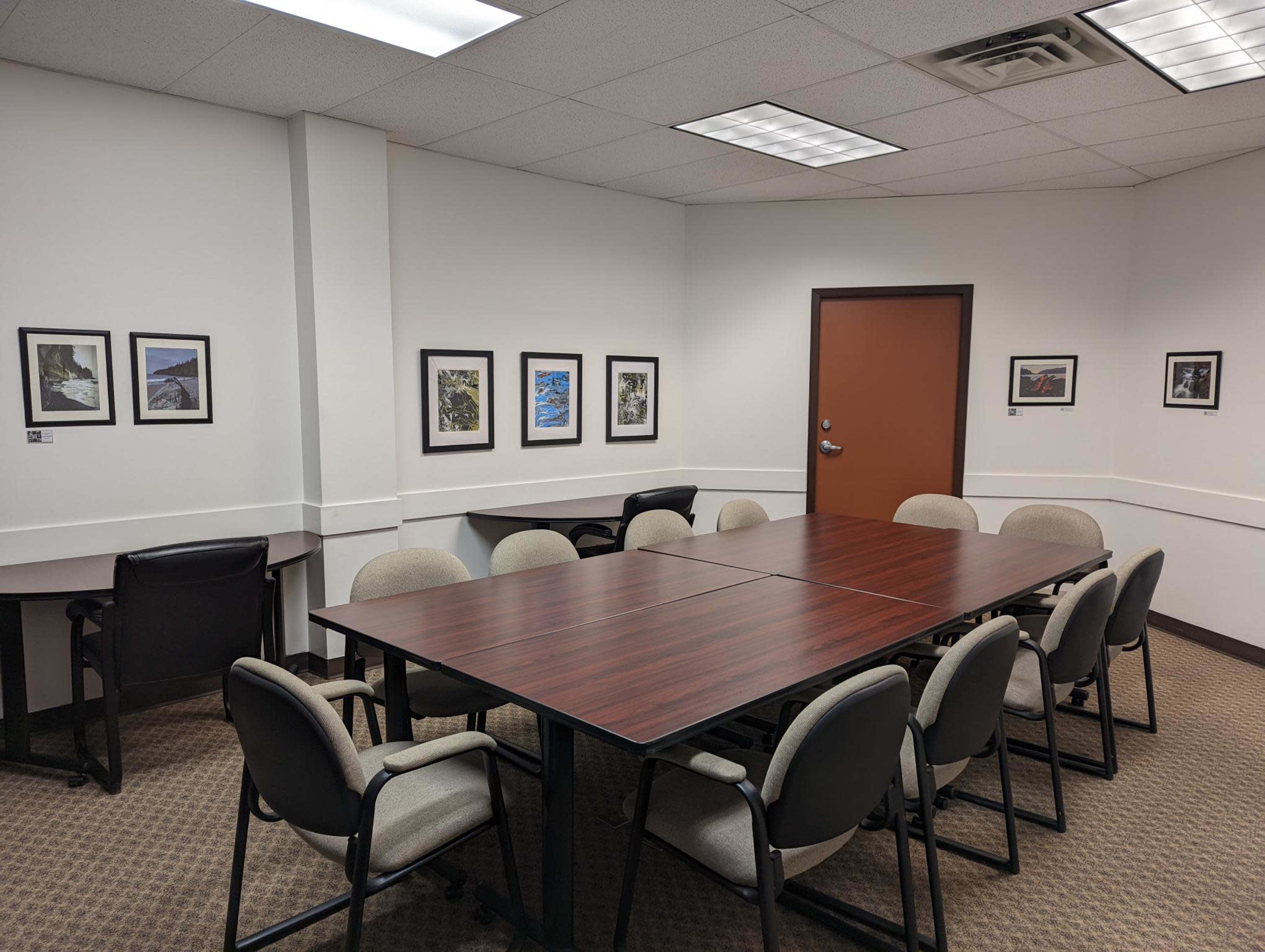 The image shows a meeting room with two large tables arranged to accommodate chairs, framed pictures on the walls, and a door leading outside.