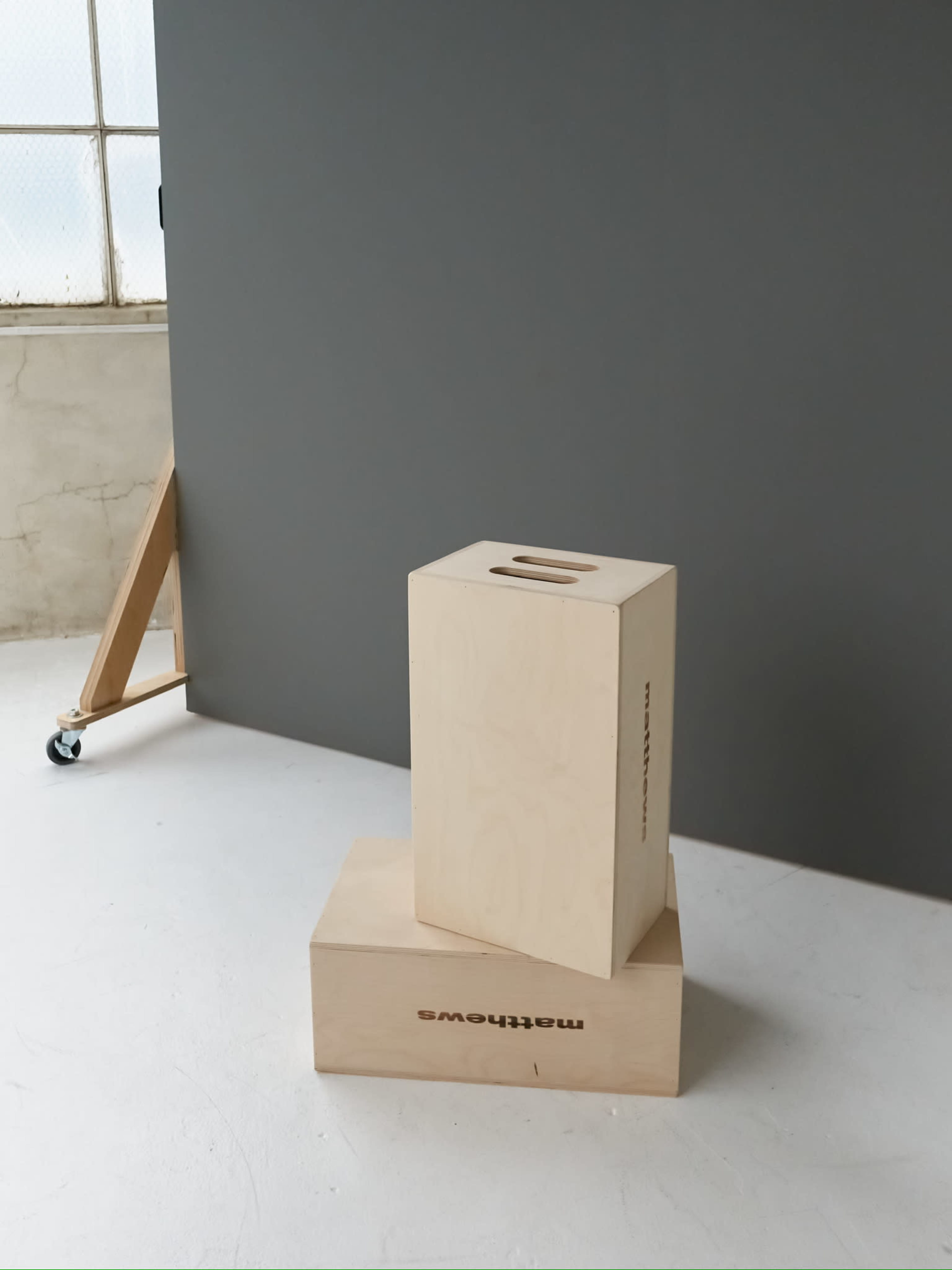 A wooden donation box is positioned on top of a larger box, set against a gray backdrop in a bright room.