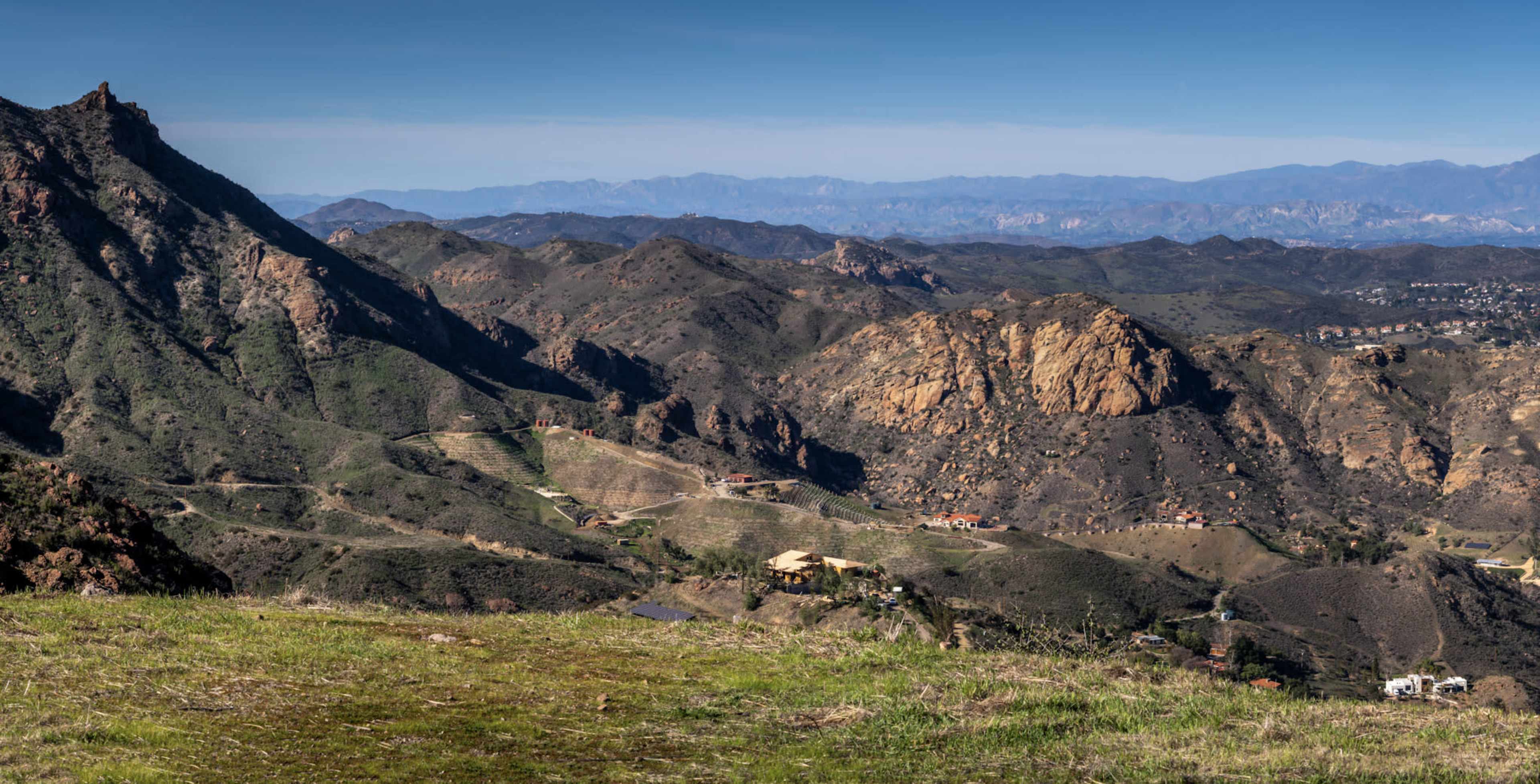 A panoramic view of rugged mountains and valleys, featuring scattered homes and rocky outcrops under a clear blue sky.