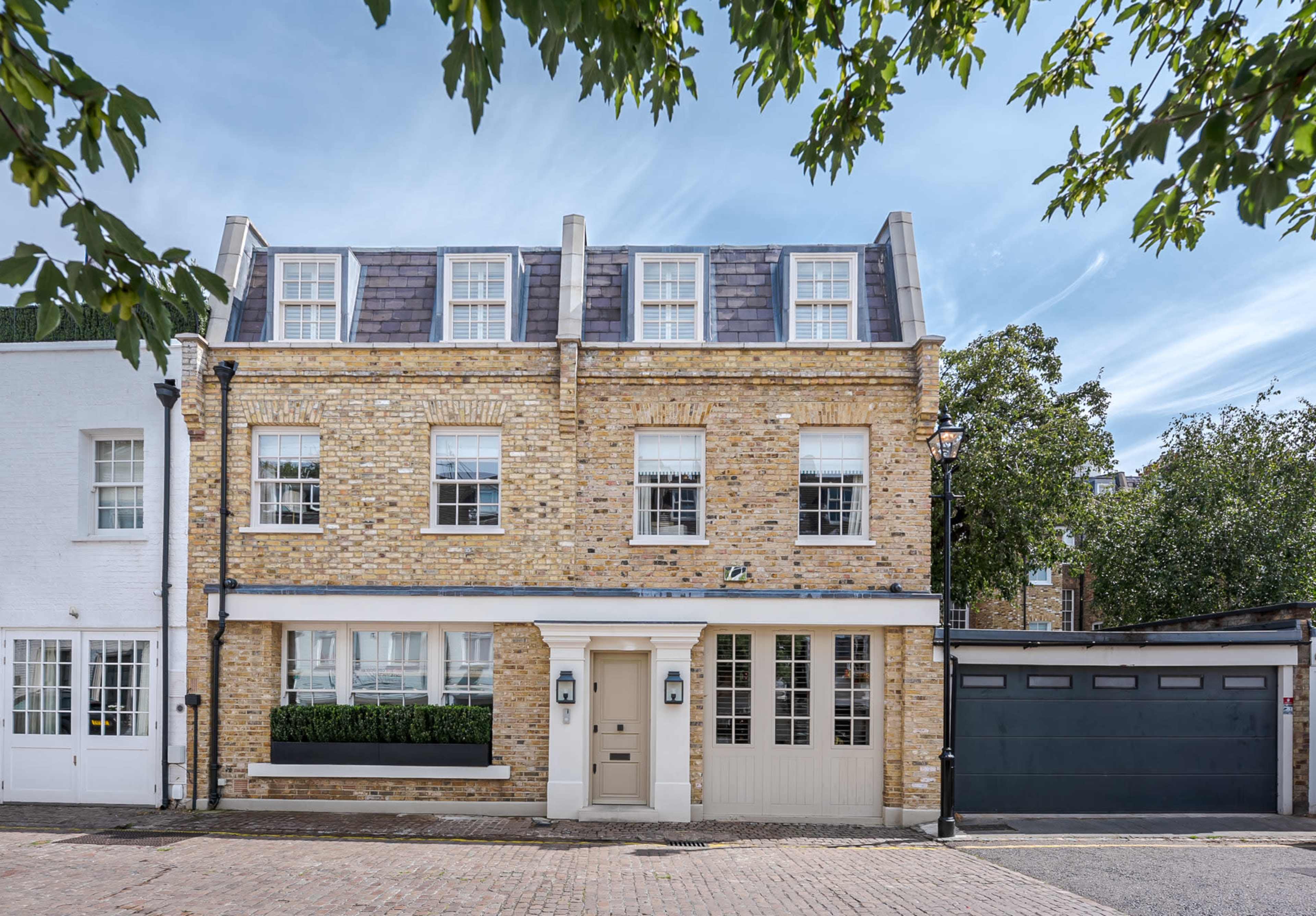 A two-story brick house with a gray roof, large windows, and a front garden, situated on a cobblestone street.