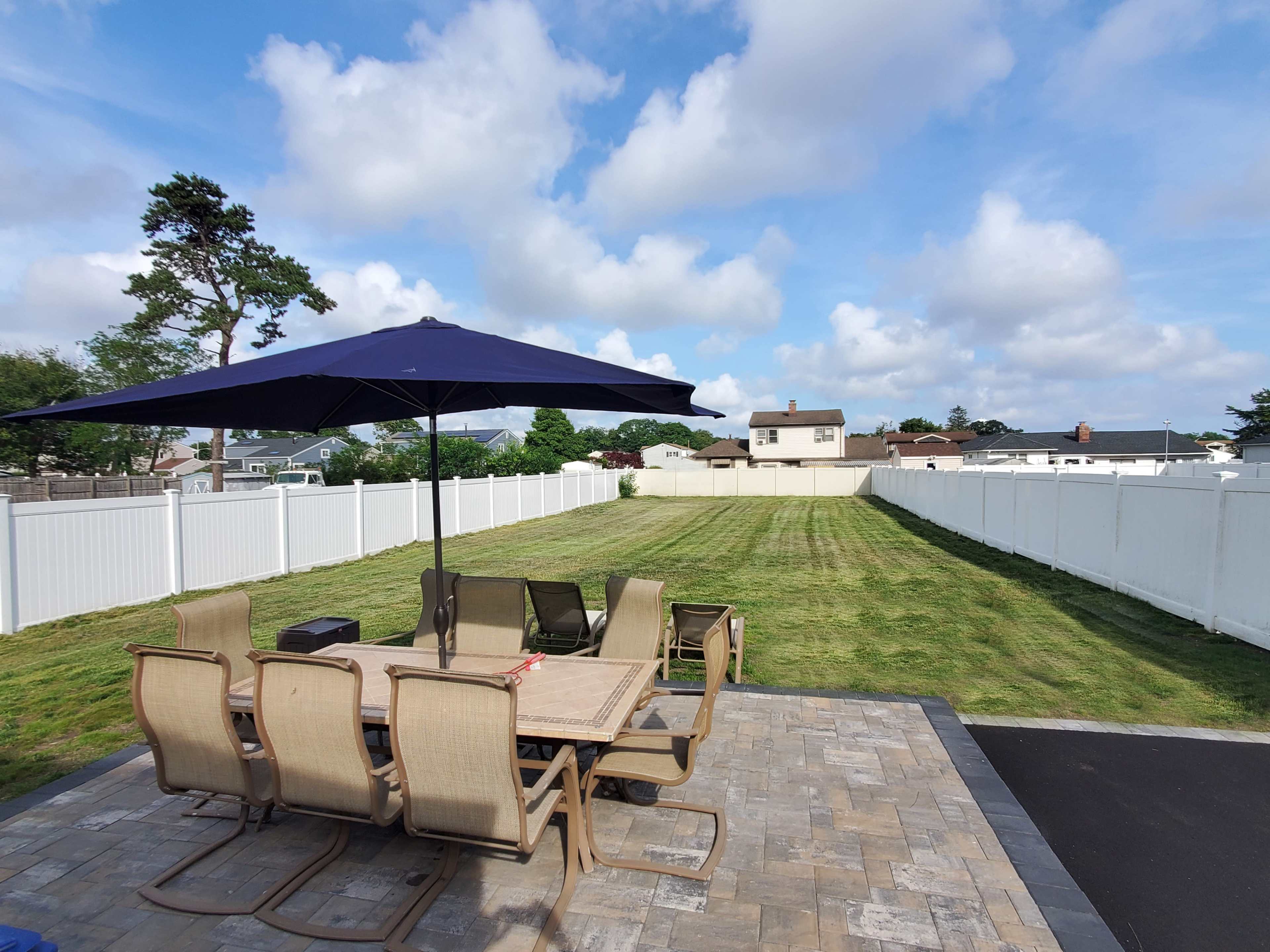 A patio set with a large umbrella overlooks a neatly mowed backyard enclosed by a white picket fence.