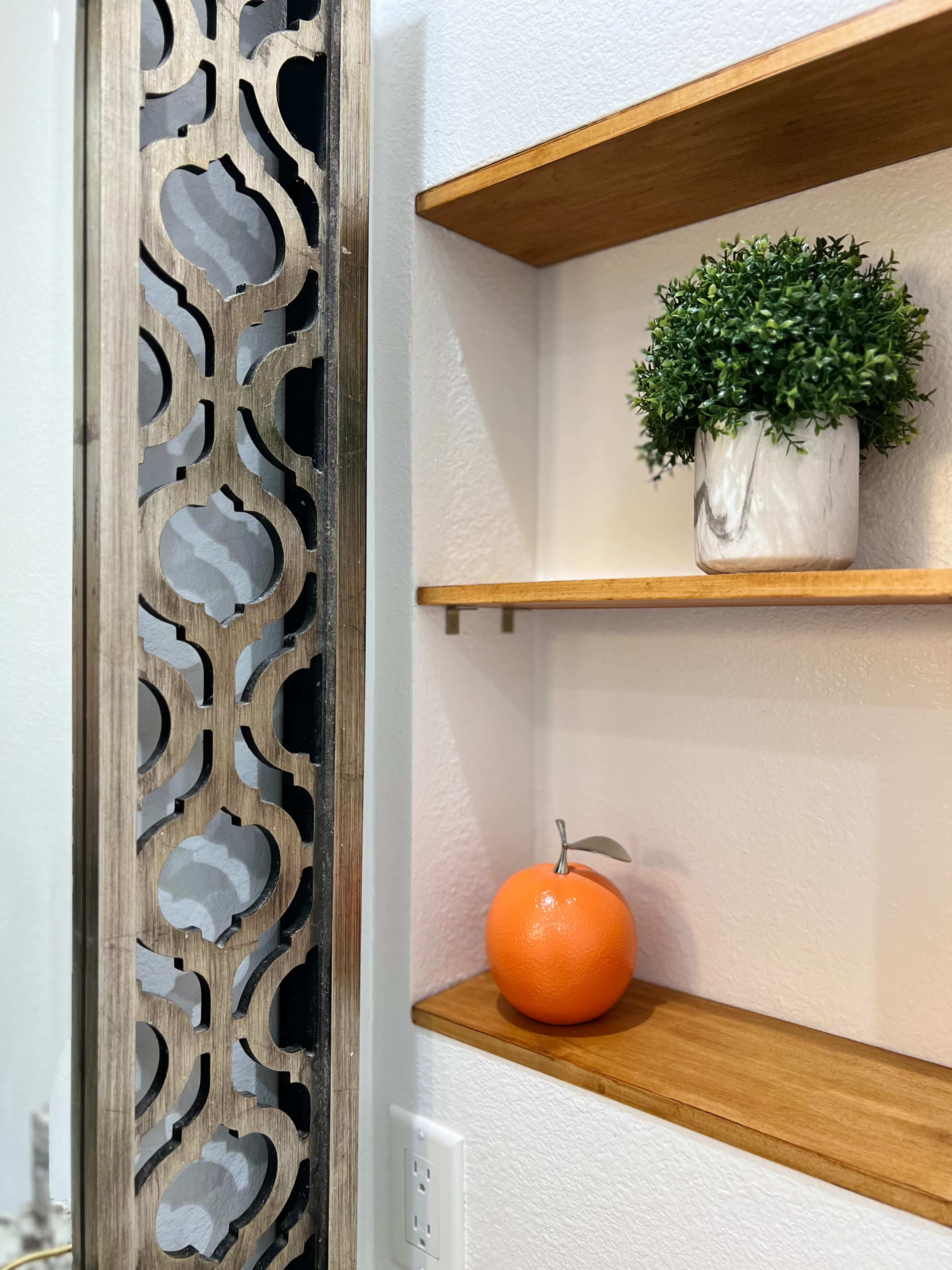 A decorative wall panel stands beside wooden shelves that hold a small potted plant and an orange.