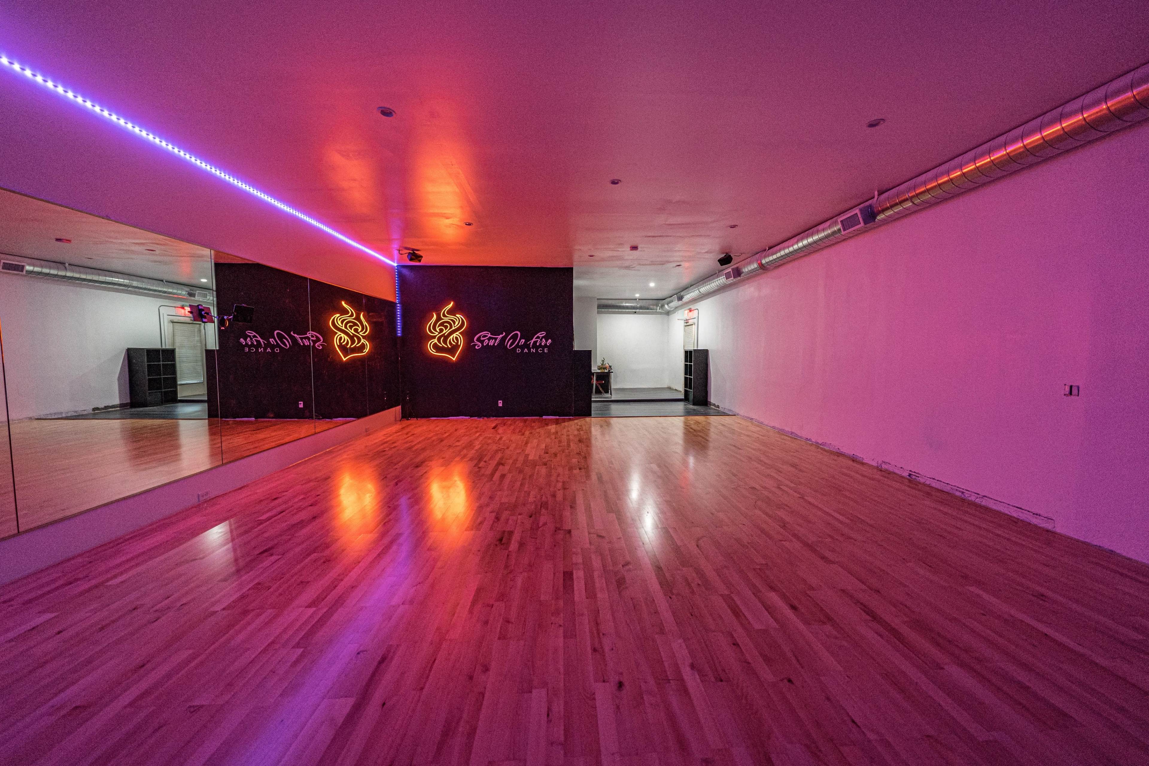 A spacious dance studio with a wooden floor, mirrored walls, and neon lighting.