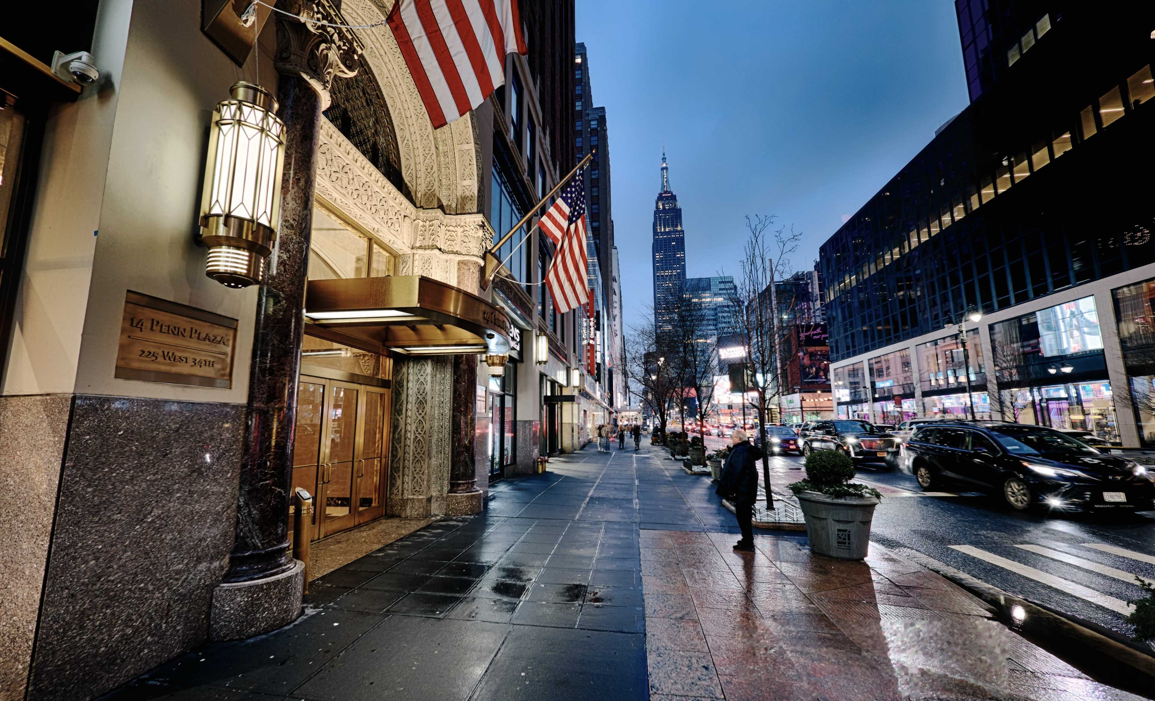 The image shows a street scene in New York City at dusk, featuring the exterior of a building with American flags, the Empire State Building in the background, and vehicles on the road.