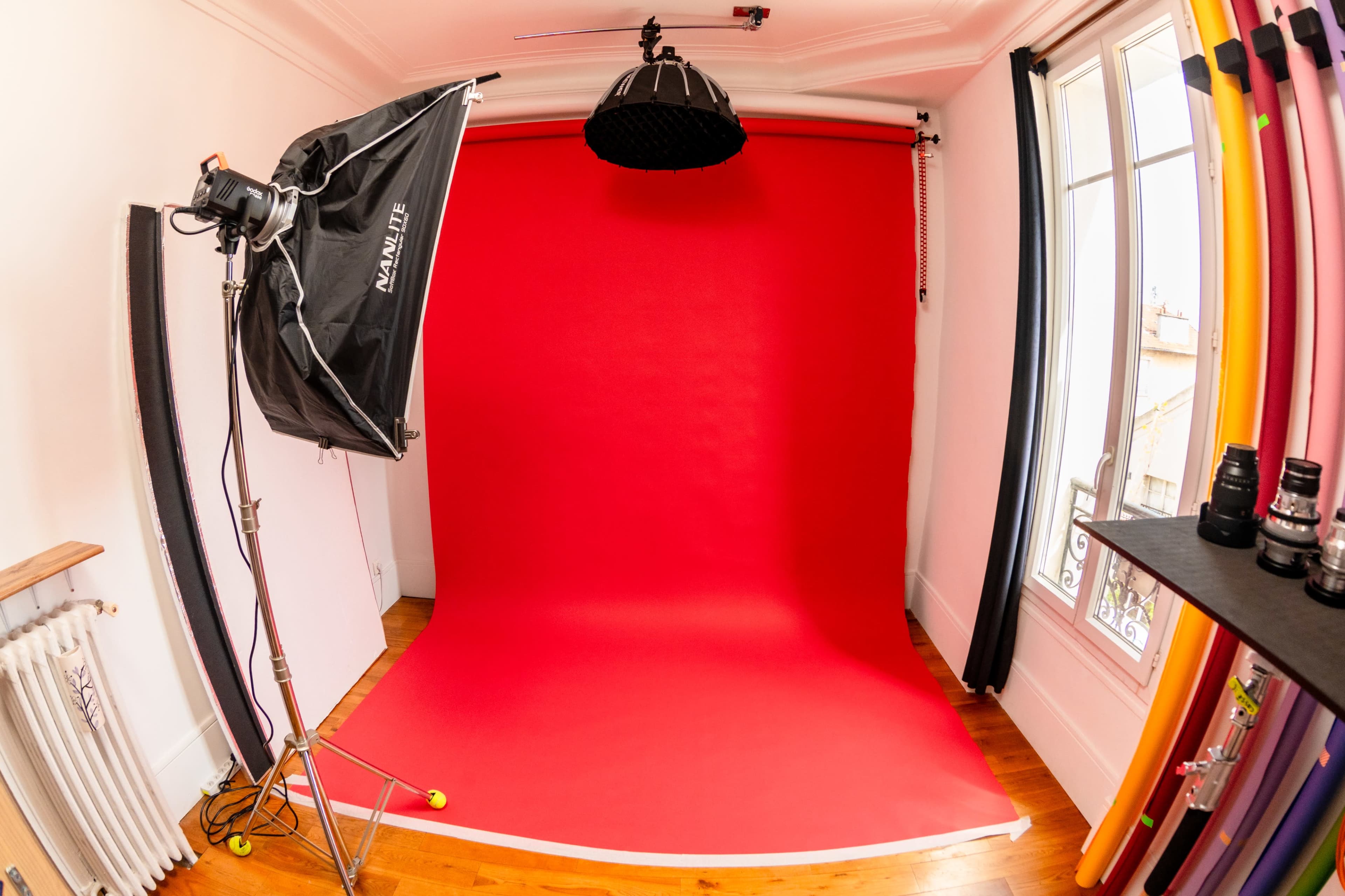 The image shows a photography studio with a red backdrop, studio lighting, and various camera lenses arranged on a shelf.