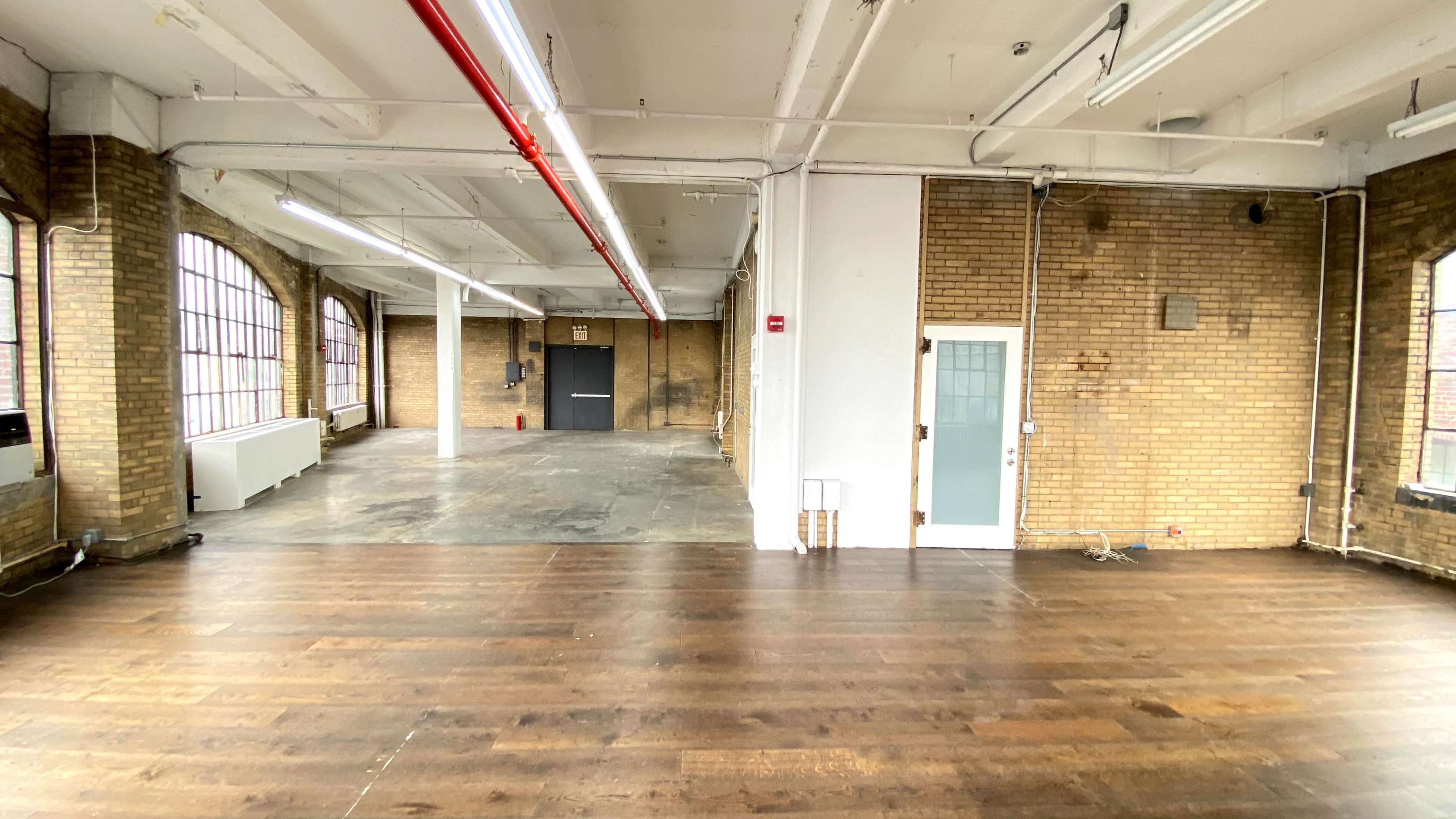 An empty industrial-style room with exposed brick walls and large windows, featuring a polished wooden floor.