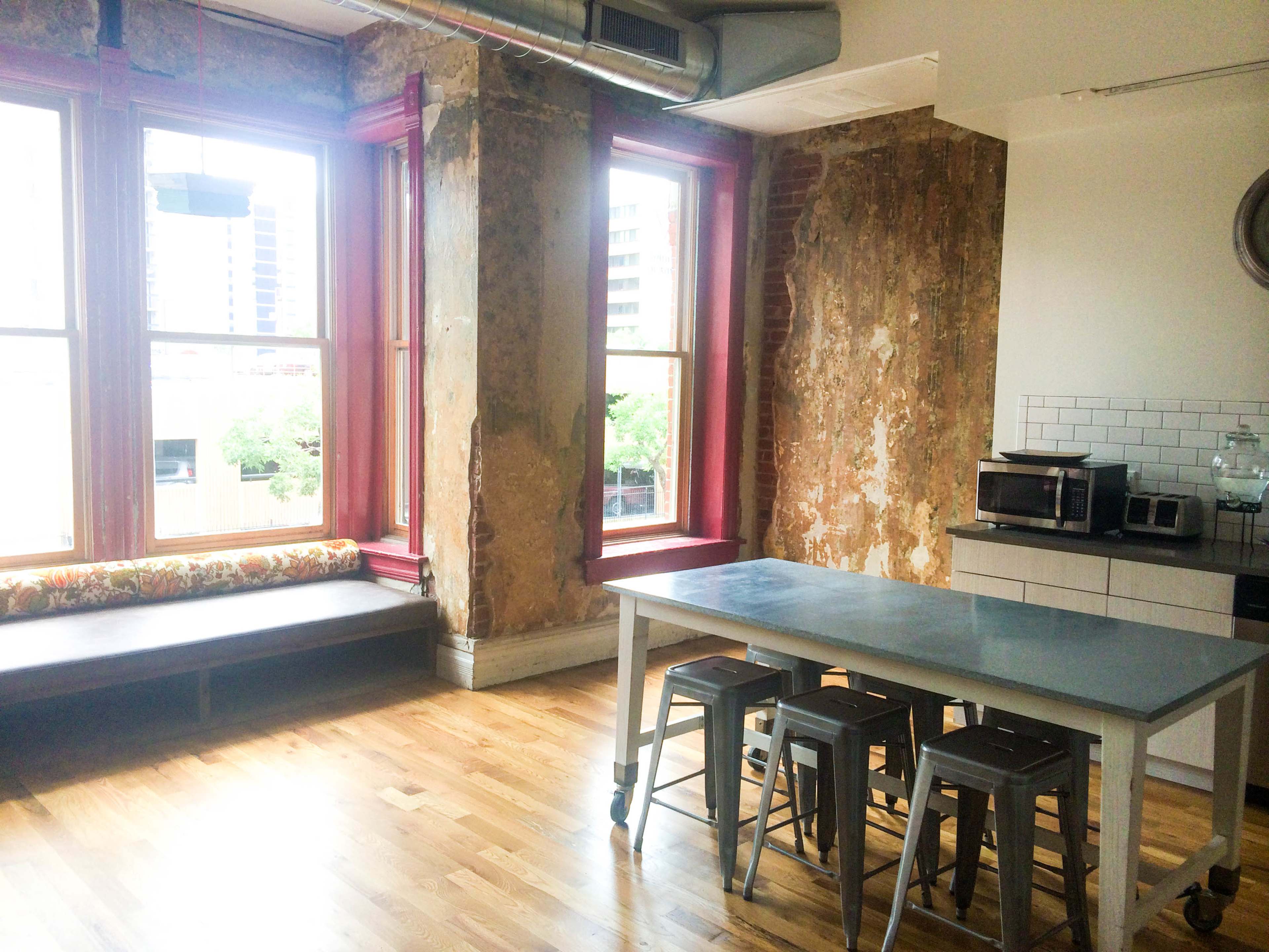A modern kitchen area with a wooden floor, steel stools, a concrete table, and large windows with red frames, surrounded by exposed brick walls.