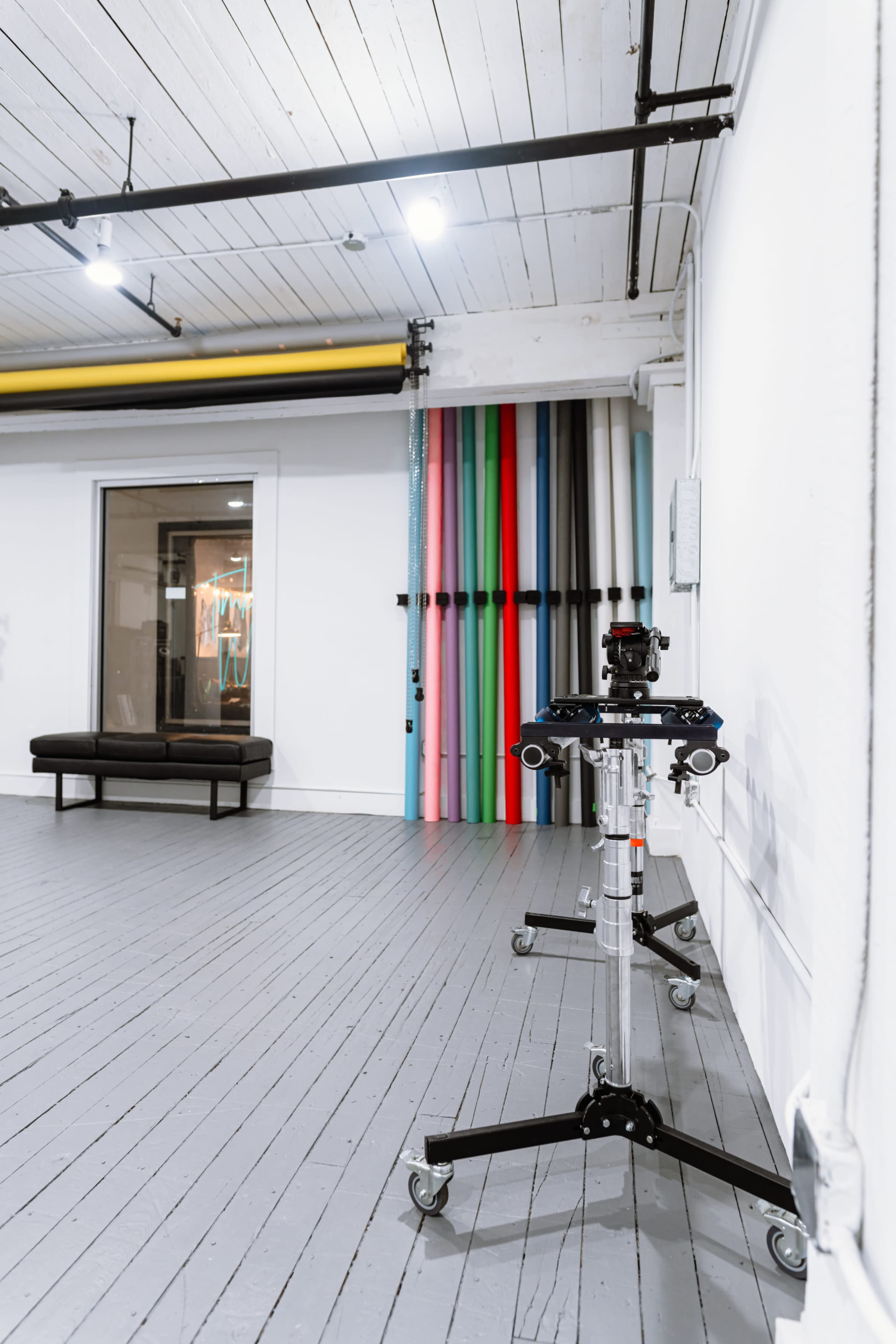 The image shows a spacious studio with a black bench, a color backdrop setup featuring vertical rolls of fabric, and a camera on a wheeled tripod in the foreground.