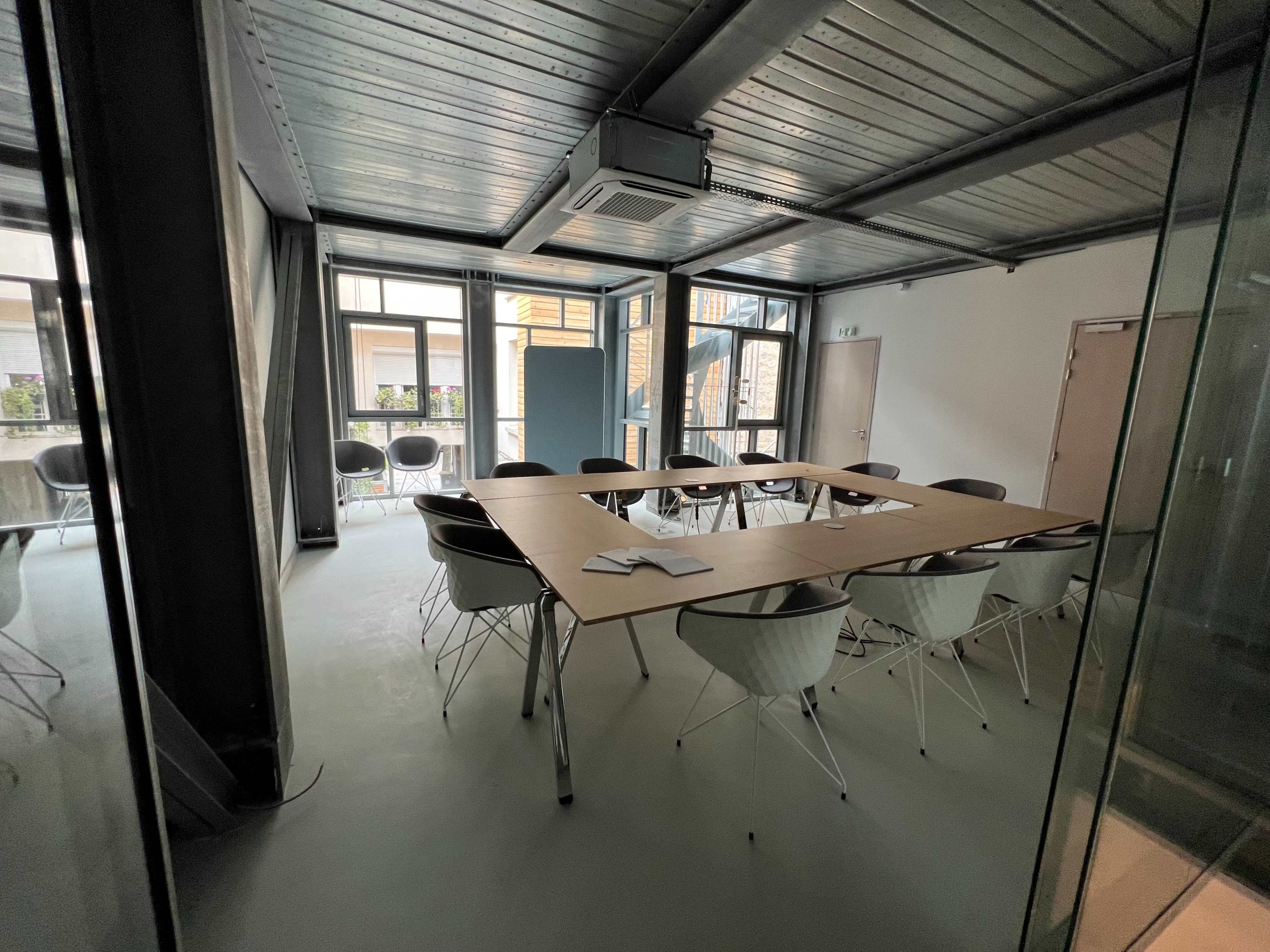 A modern conference room with a large rectangular table surrounded by white chairs, featuring windows that provide natural light.
