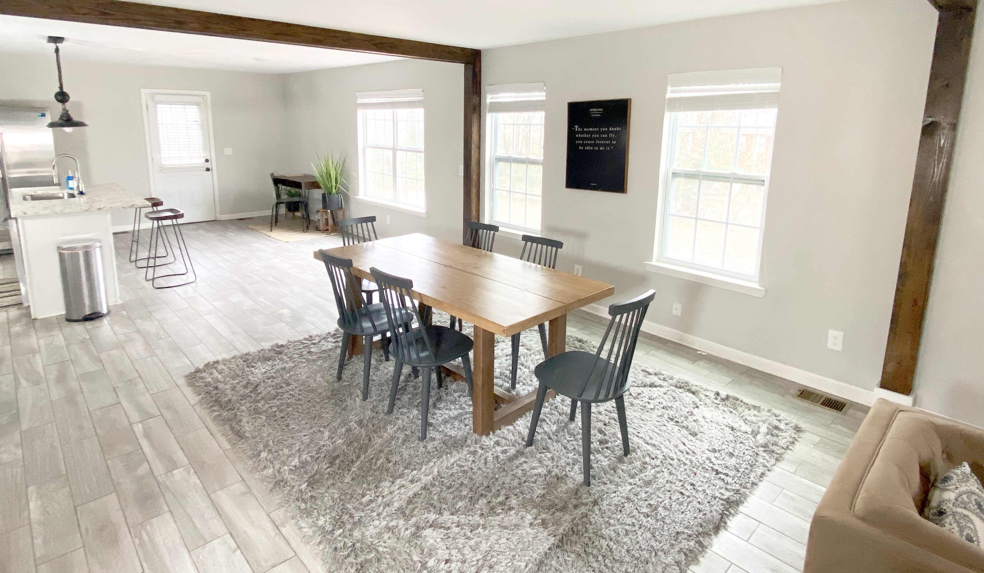 A dining area features a wooden table surrounded by chairs on a plush rug, with large windows letting in natural light.