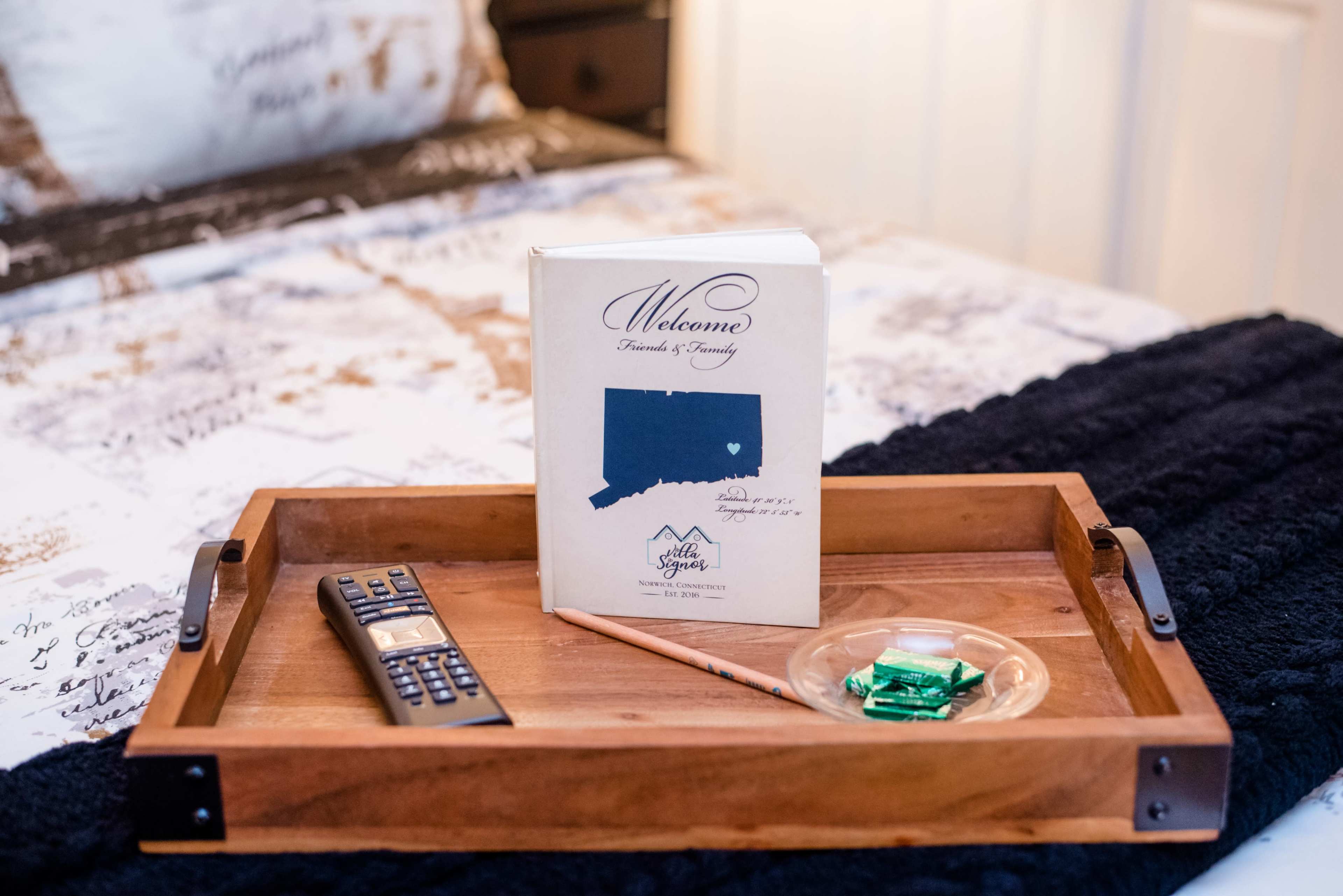 A wooden tray on a bed holds a welcome book, a remote control, a pencil, and a dish with green candies.