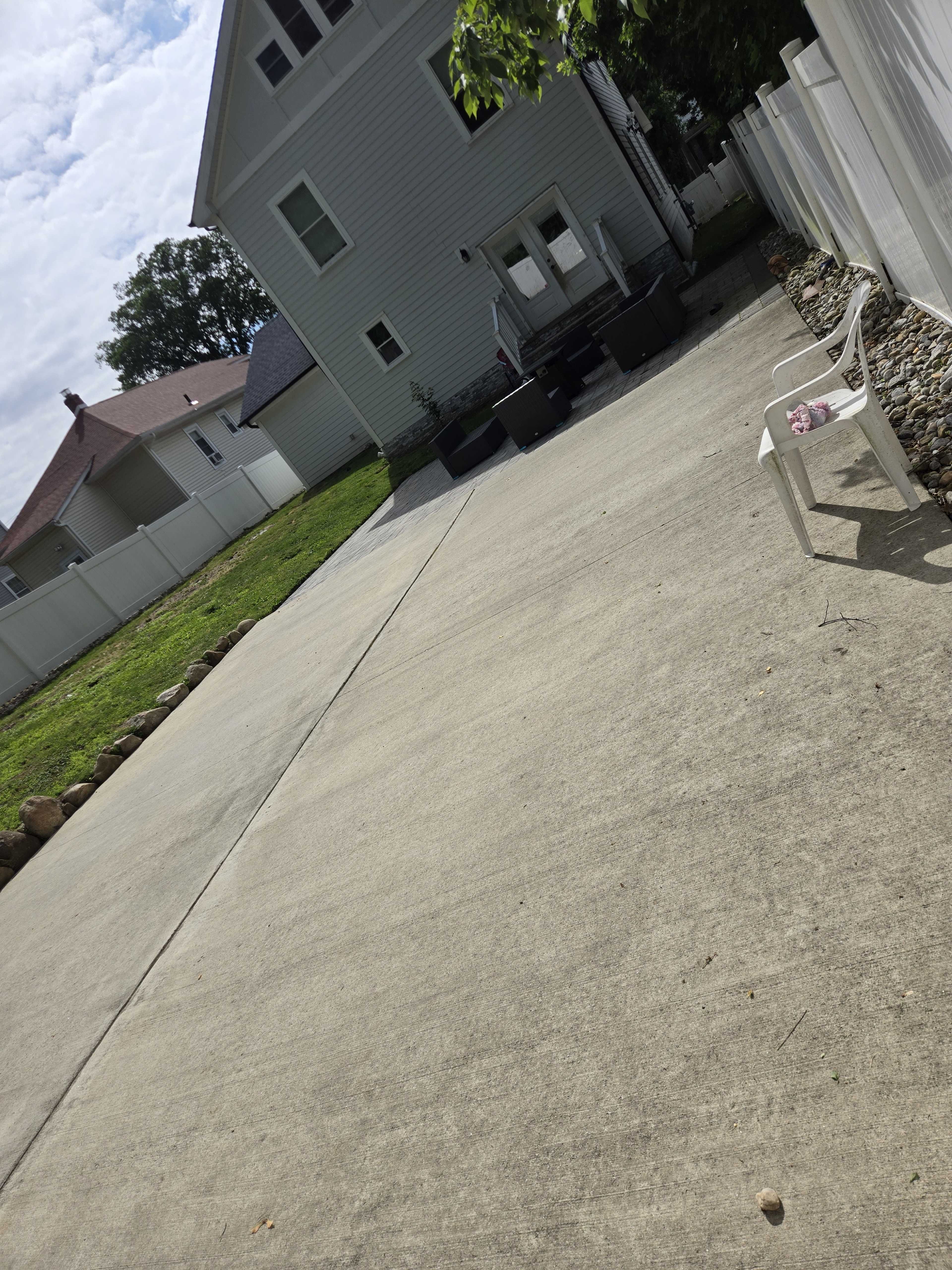 The image shows a concrete driveway leading to a house with a fenced yard, bordered by rocks and a single white chair on one side.