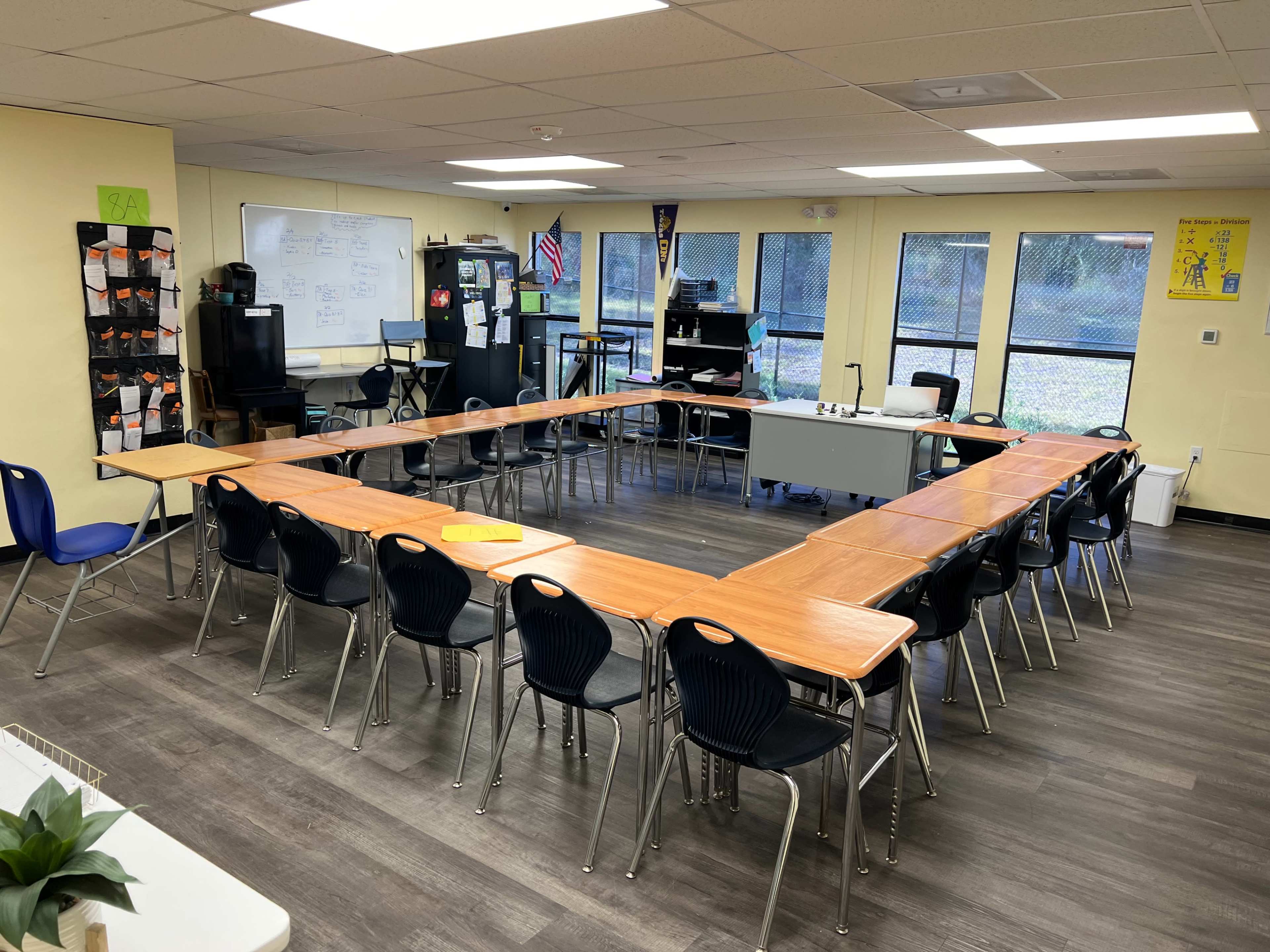 A classroom arranged with several tables in a U-shape, surrounded by black chairs, with windows and a whiteboard in the background.