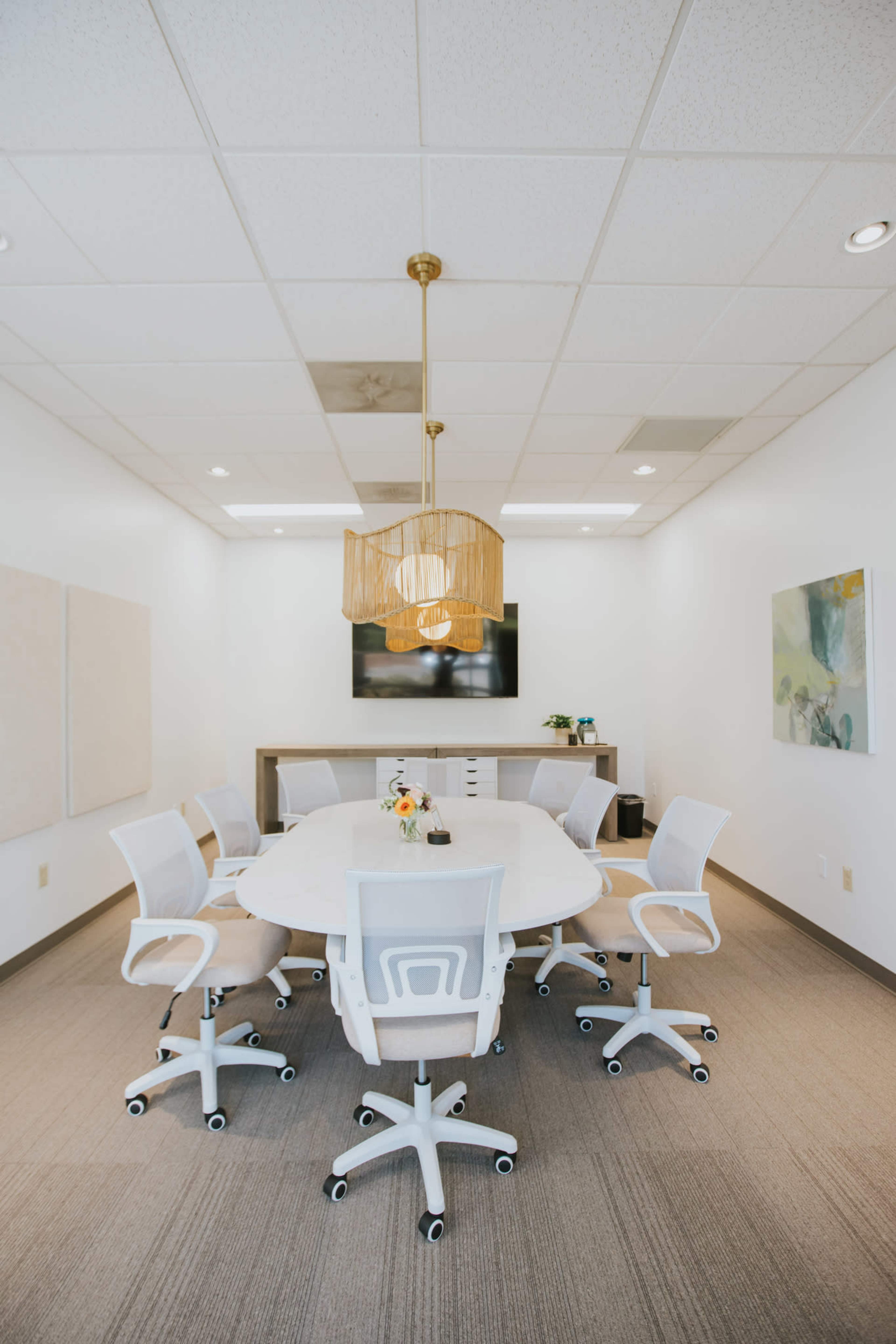 A modern conference room features a large oval table surrounded by white chairs, a decorative light fixture, and a wall-mounted television.