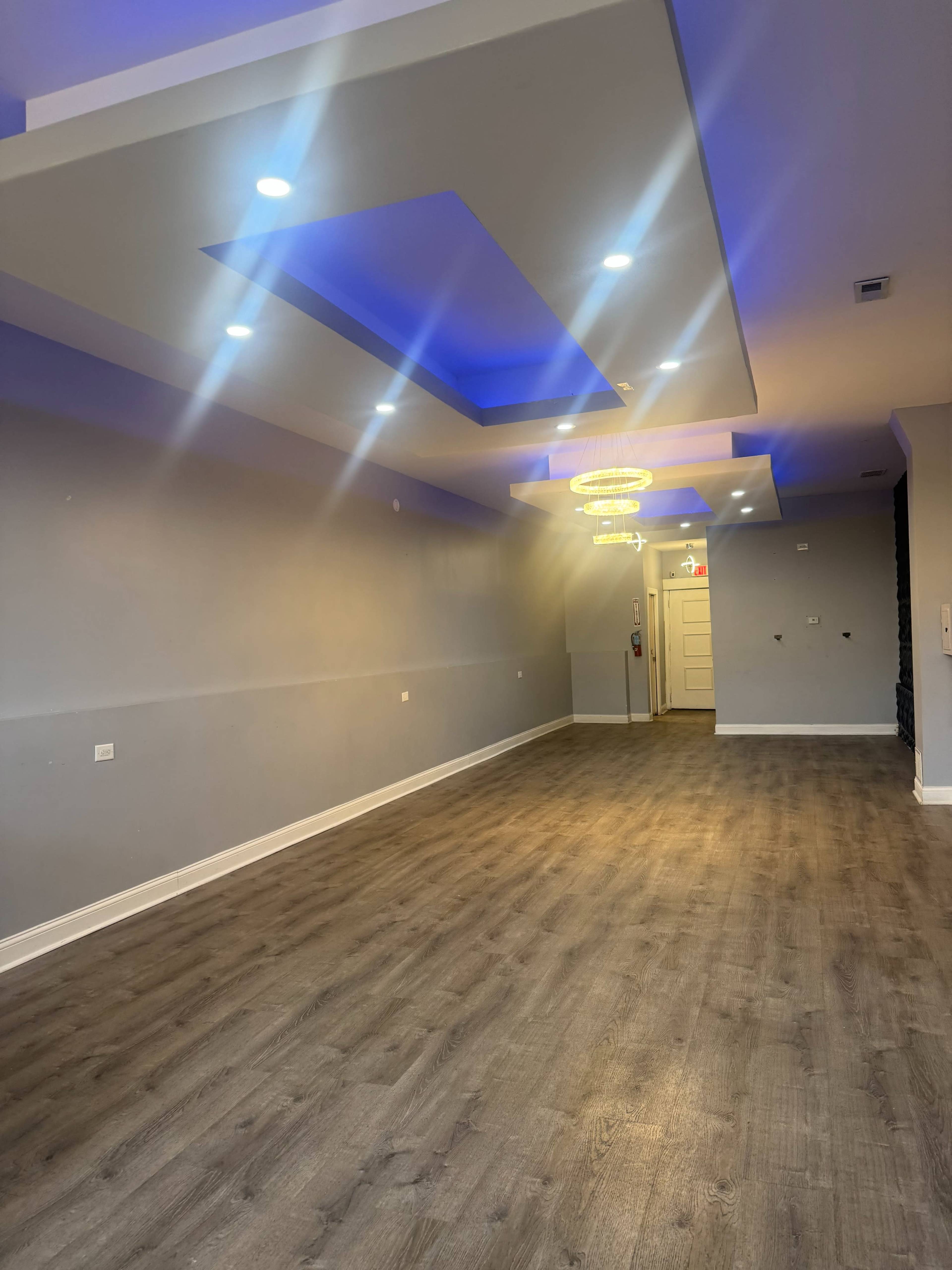 The image shows a spacious room with gray walls, modern ceiling lighting, and a chandelier, featuring hardwood flooring.
