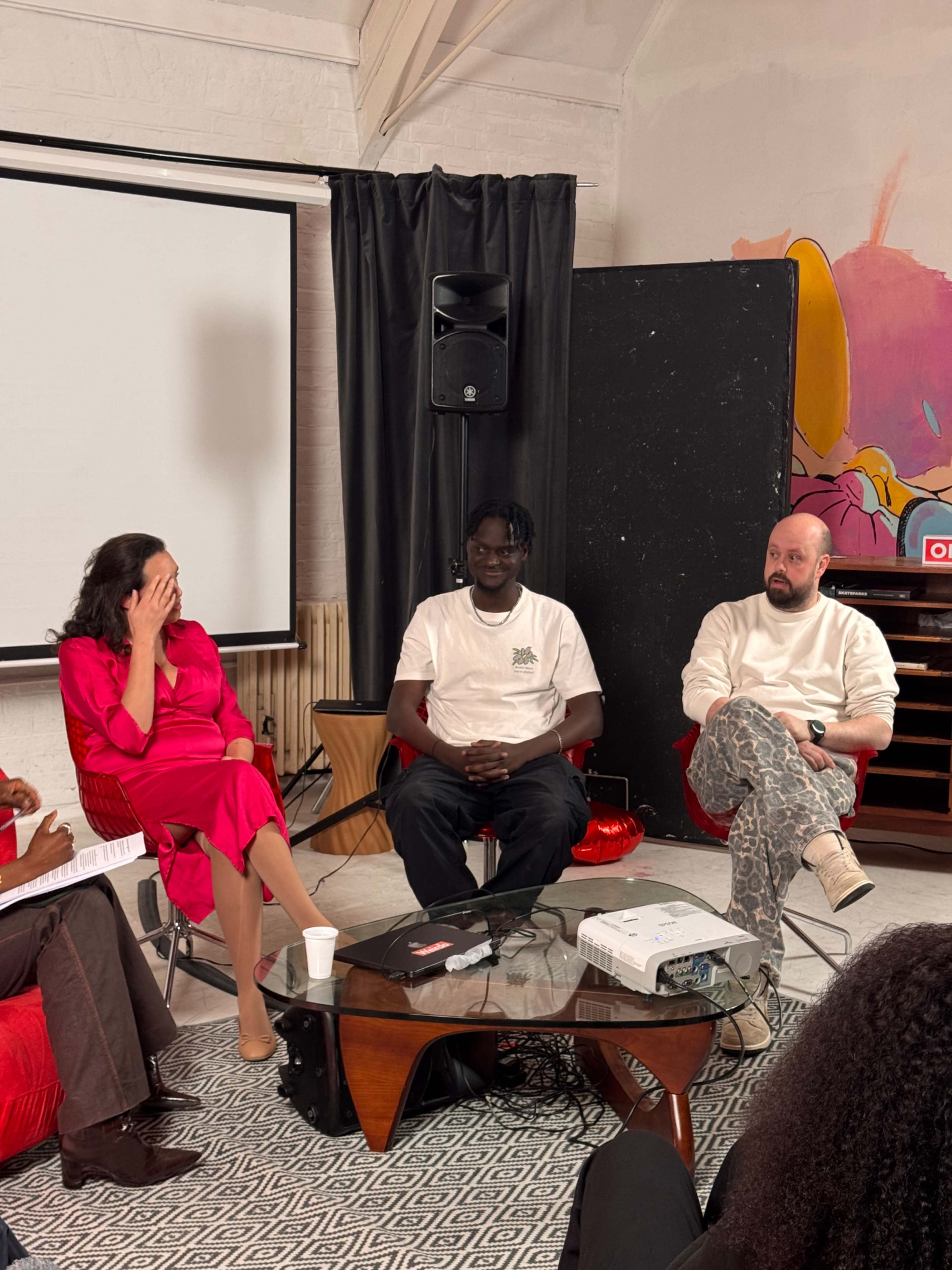 Three individuals are seated in a modern discussion setting, with a projector and a screen behind them, while a fourth person, partially visible, seems to be taking notes.