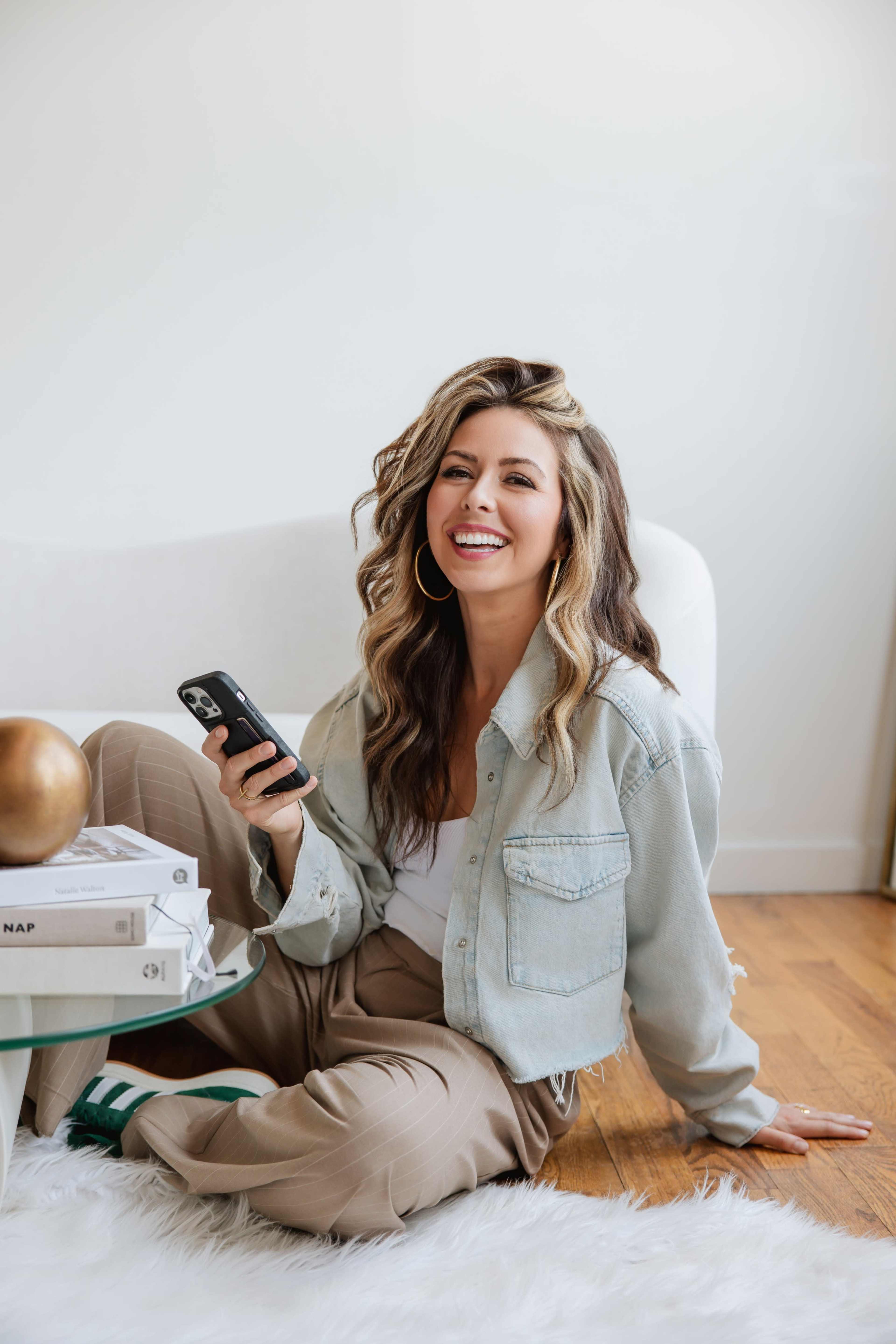 A woman with wavy hair sits on the floor next to a glass table, holding a phone and smiling while wearing a denim jacket and beige pants.