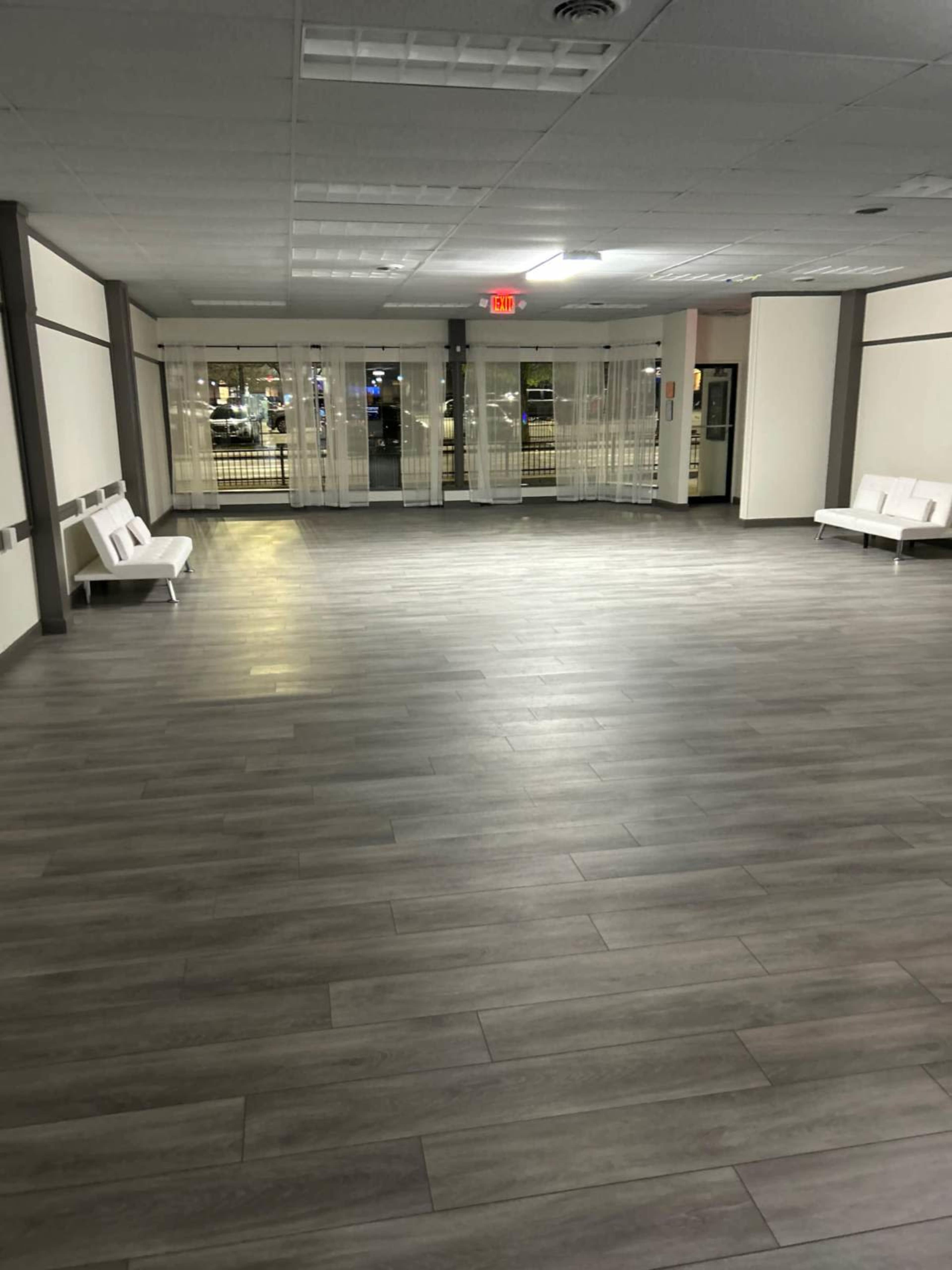 The image shows an empty room with gray flooring and white benches, illuminated by overhead lights and featuring large windows with sheer curtains.