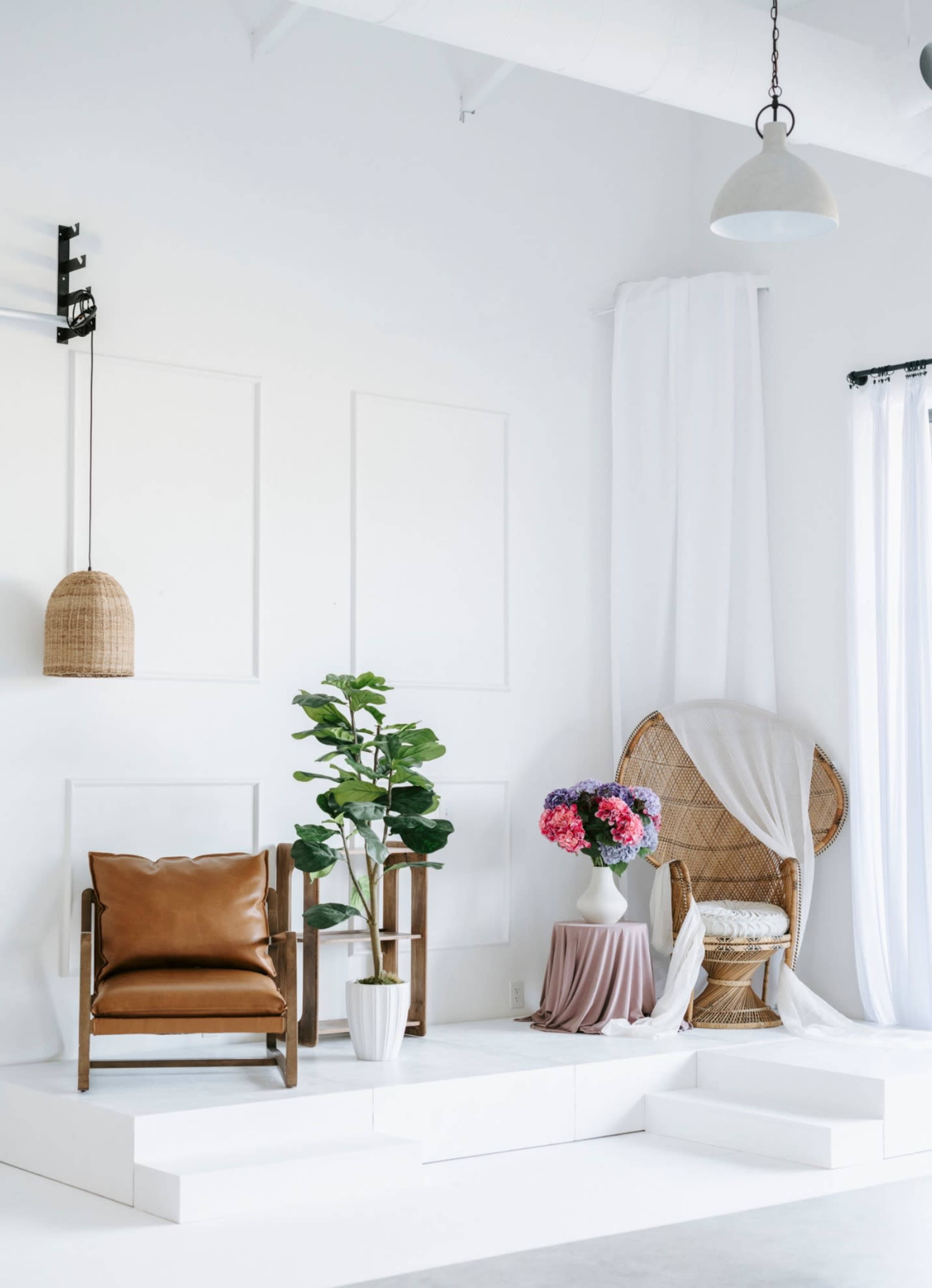 The image shows a minimalist interior scene featuring a brown leather chair, a large potted plant, a rattan chair adorned with white drapery, and a vase of colorful flowers on a pedestal.