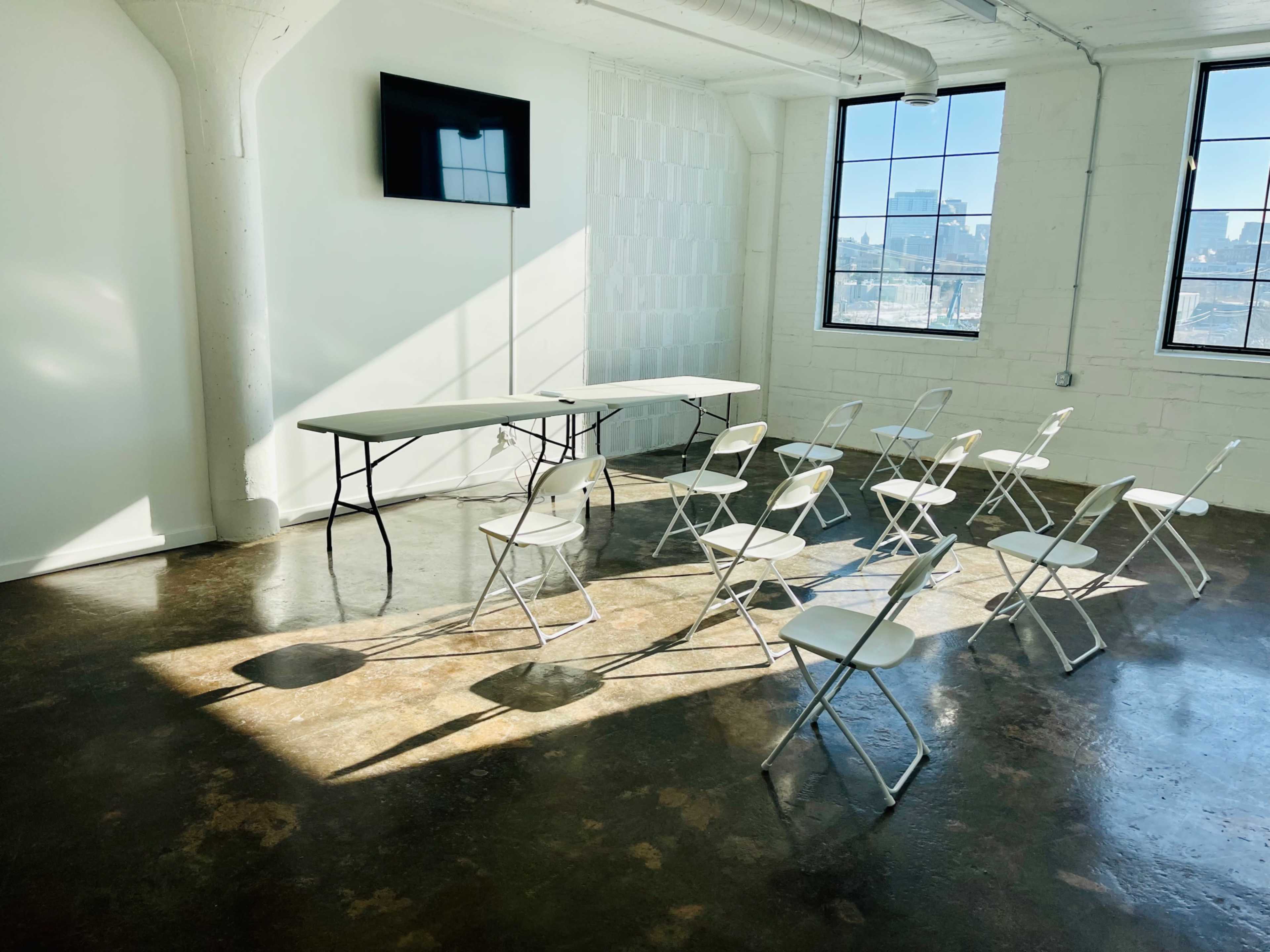 A bright, newly furnished meeting room features a rectangular table and several folding chairs arranged in front of a wall-mounted screen.