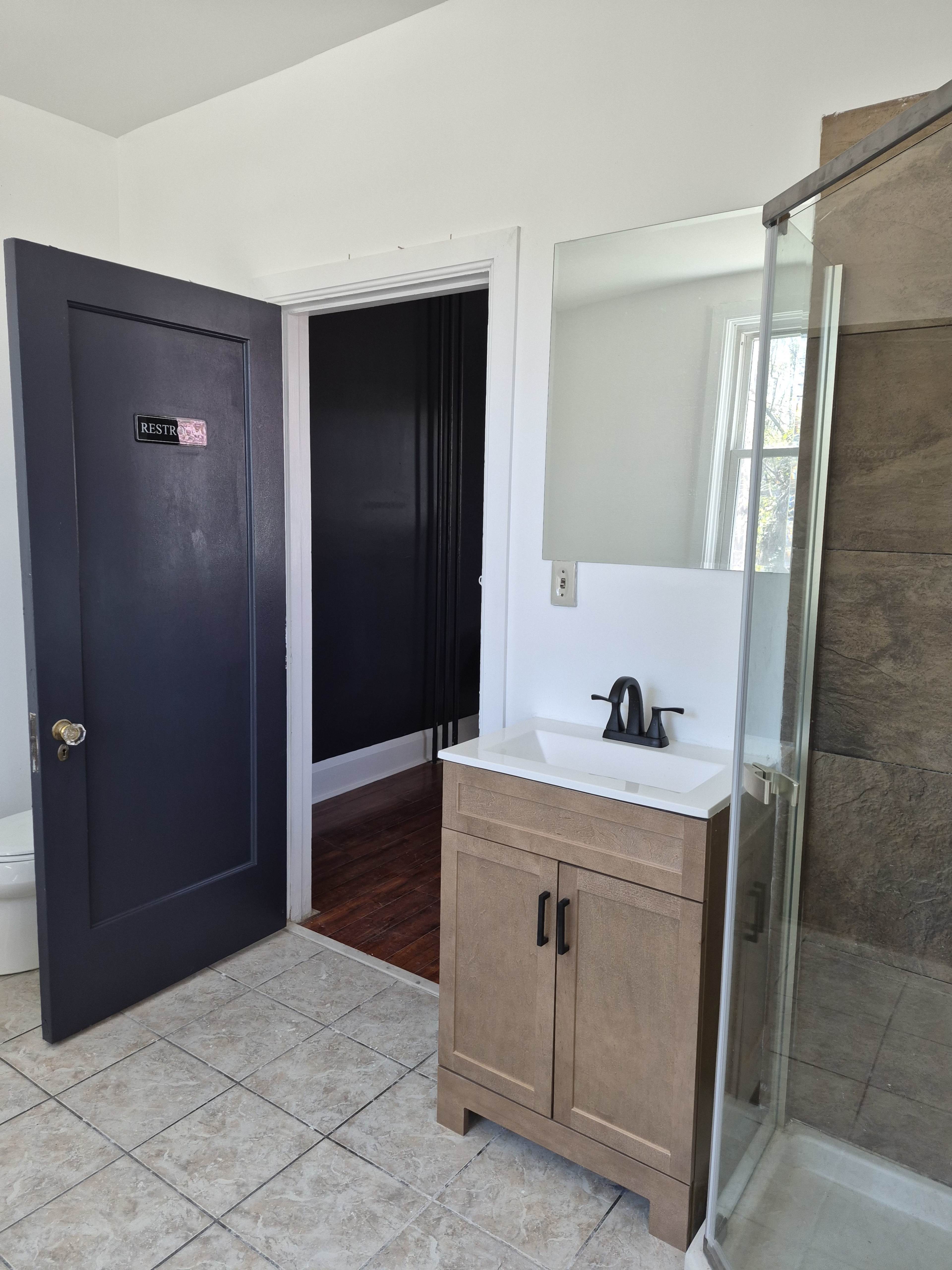The image shows a bathroom featuring a wooden vanity with a sink, a glass shower unit, and a doorway leading to a dark-painted room.
