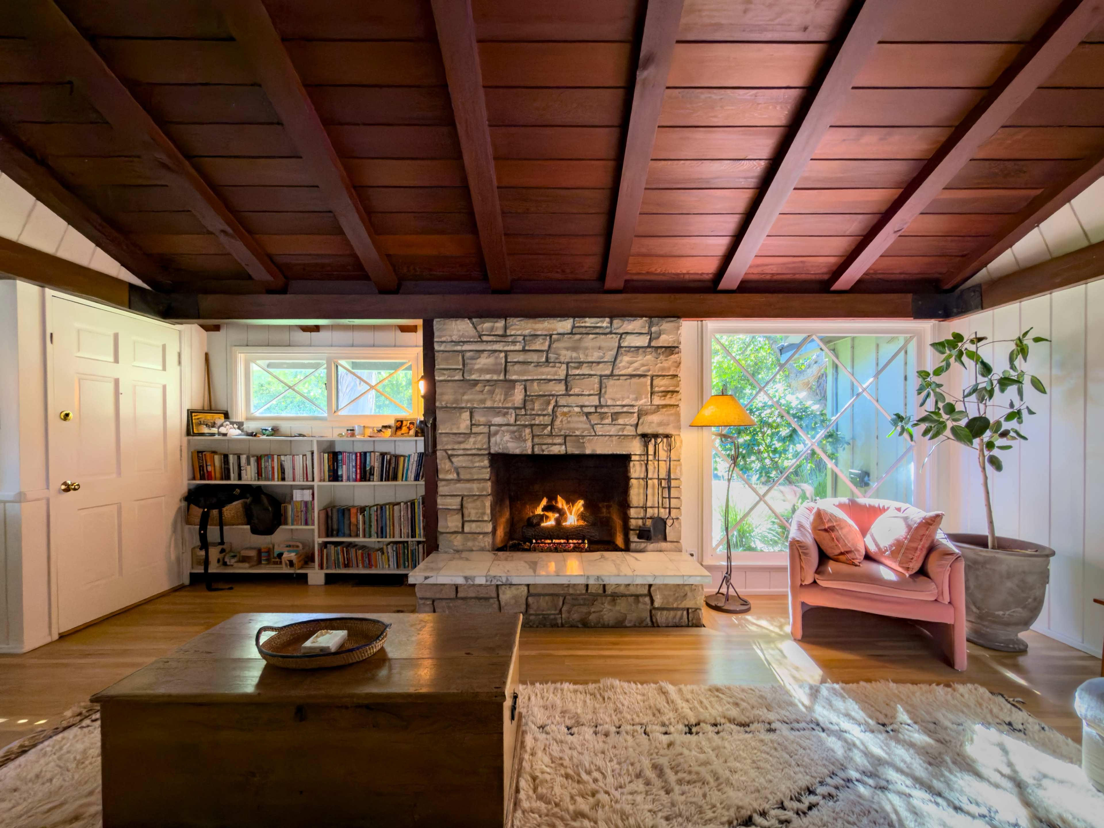 The image shows a cozy living room with a stone fireplace, wooden ceiling beams, bookshelves, and a pink armchair next to a window.