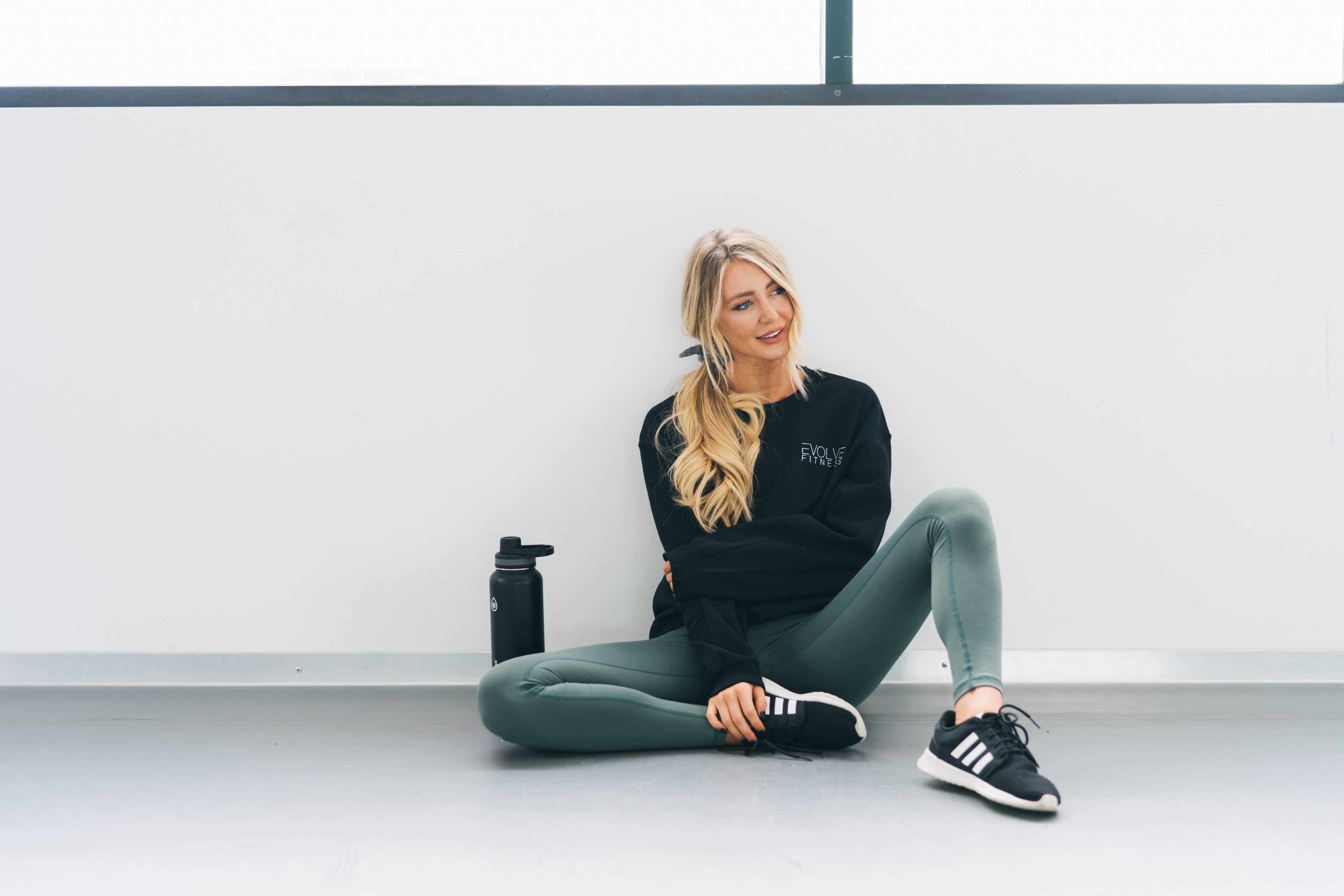 A woman is sitting on the floor in a gym, wearing black and green athletic clothing with a water bottle beside her.