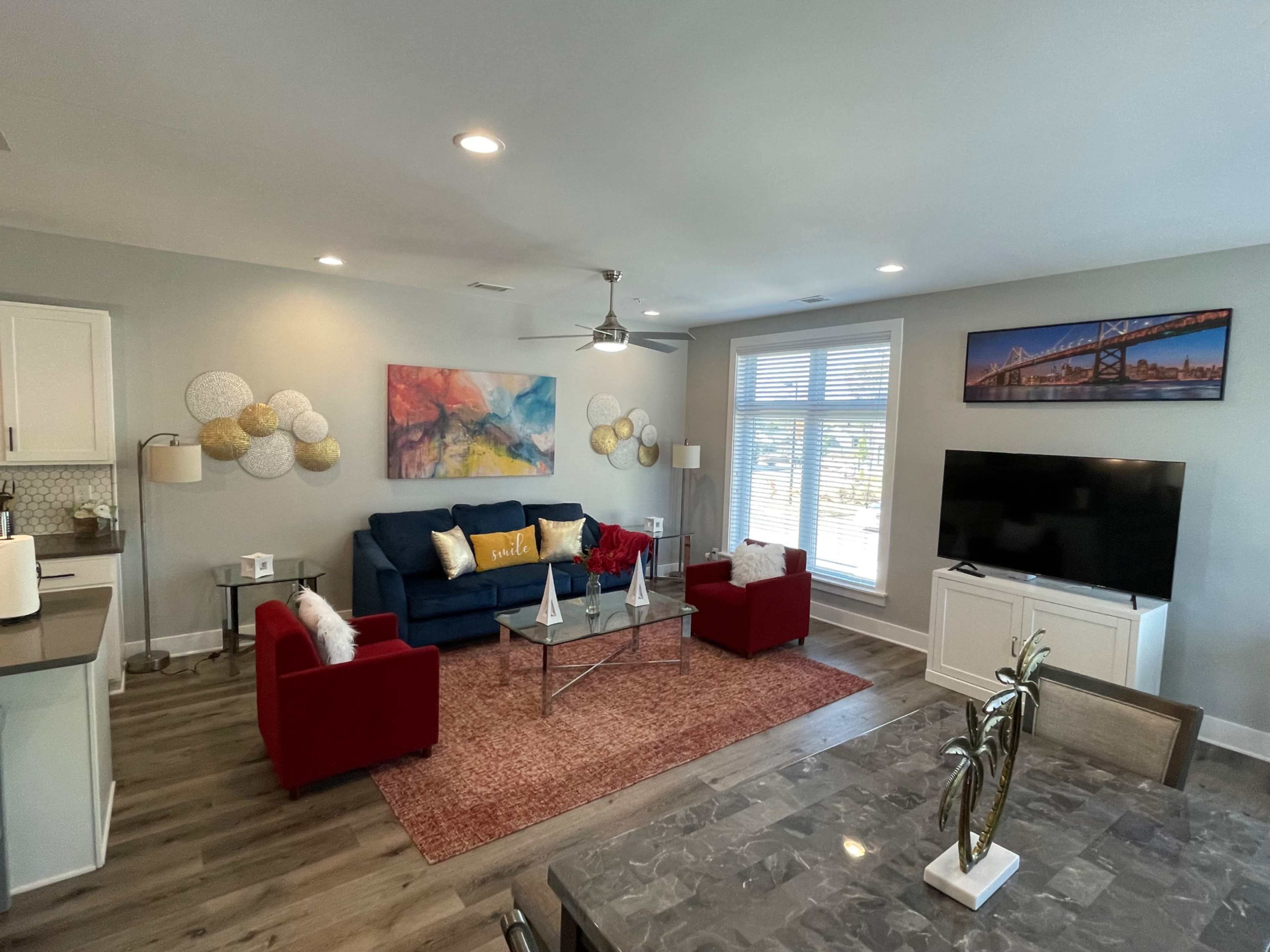 The image shows a modern living room with a blue sofa, red chairs, a coffee table, and a large television, all illuminated by natural light from a nearby window.