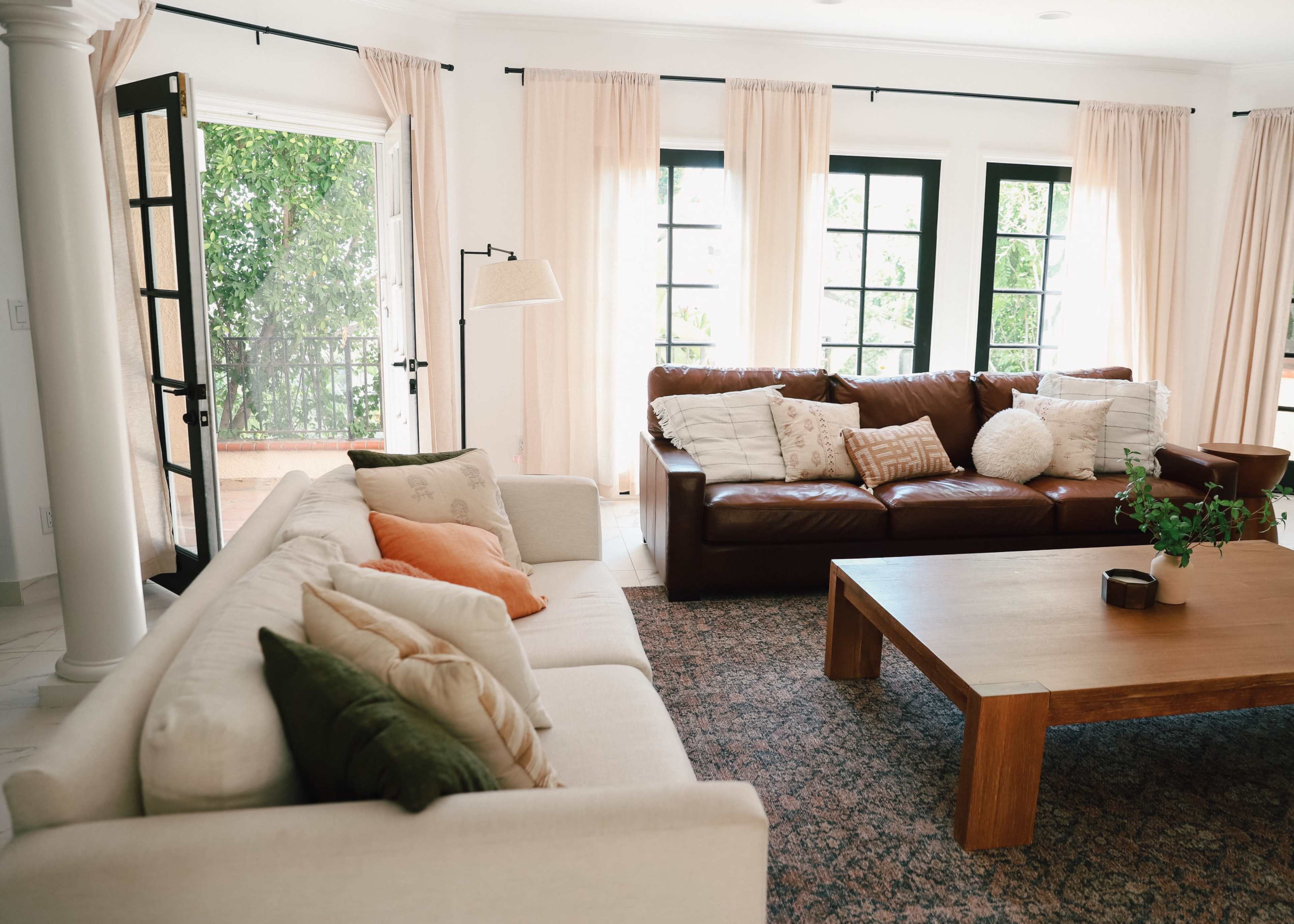 A living room features a light-colored sofa and a brown leather couch facing a wooden coffee table, with large windows draped in cream curtains.