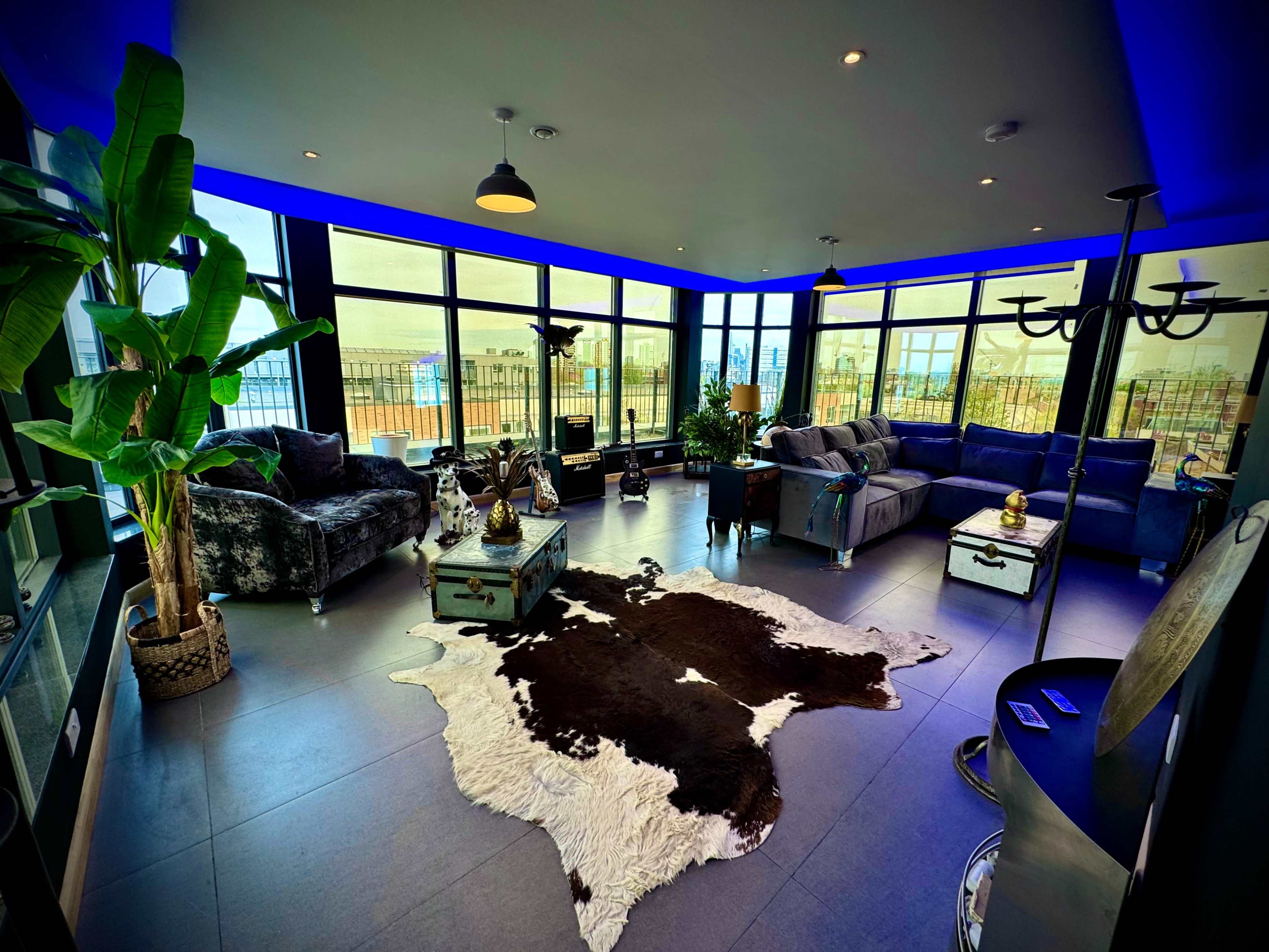 The image shows a modern living room with large windows, blue ambient lighting, several seating areas, and a cowhide rug on the floor.