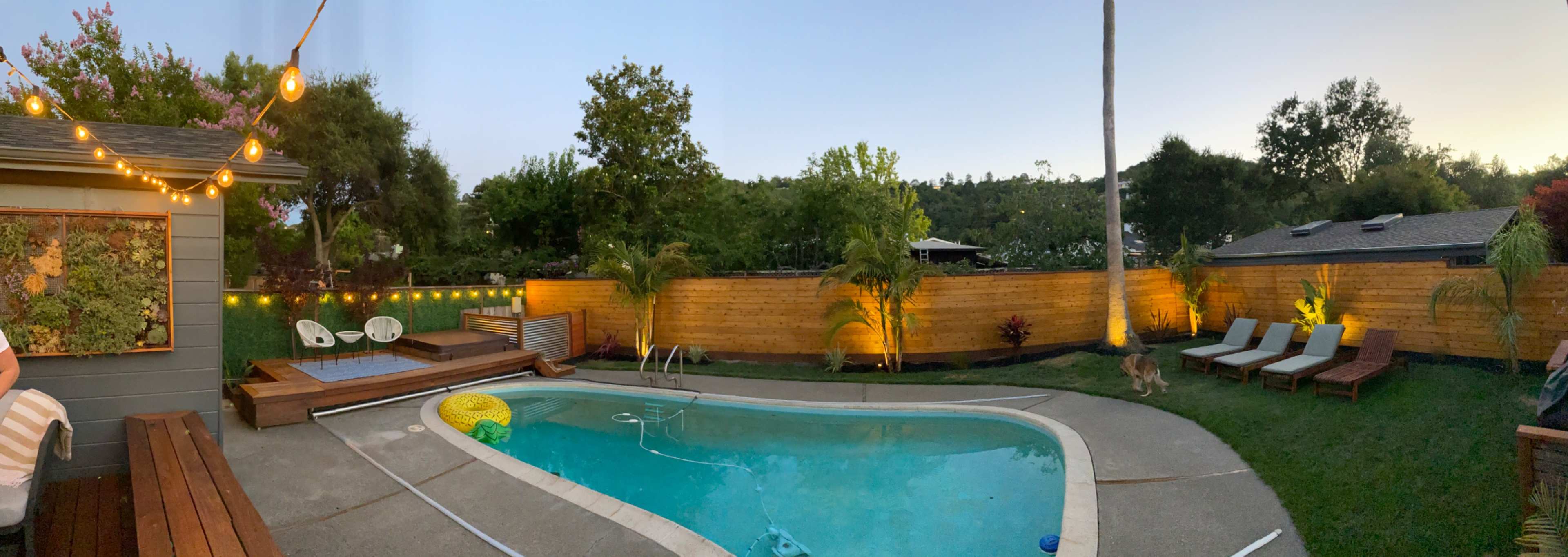 A backyard with a swimming pool, lounge chairs, and string lights illuminating a surrounding landscaped area in the evening.