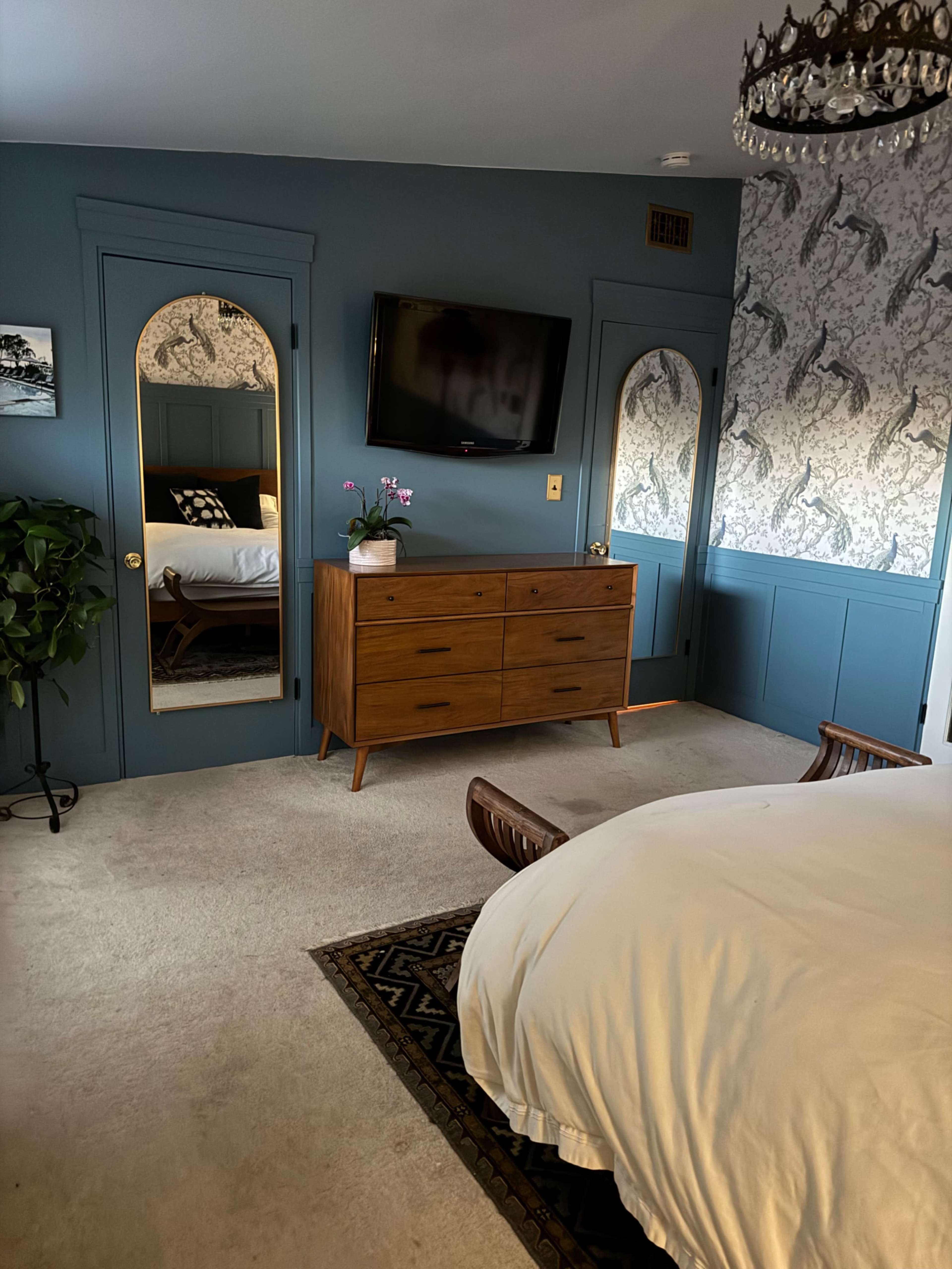 The image shows a neatly arranged bedroom featuring a wooden dresser, a wall-mounted TV, and decorative mirrors against a blue wall, with a bed partially visible.