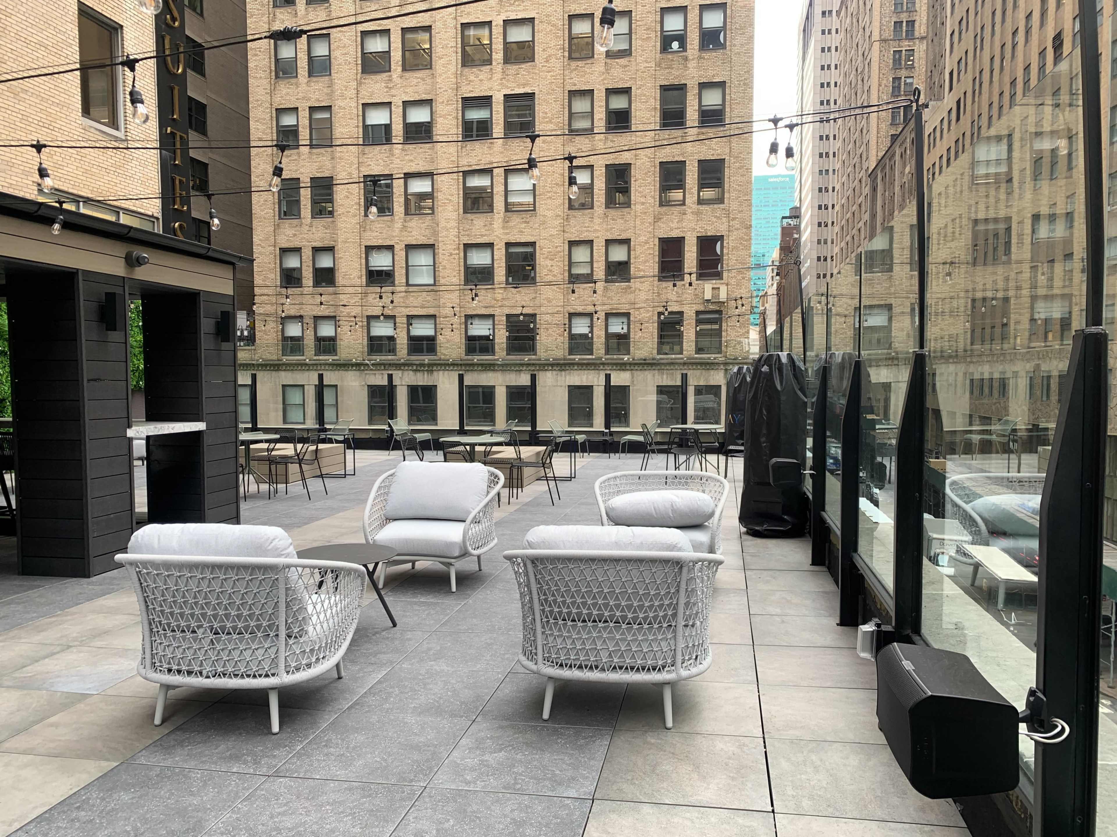 The image shows a rooftop terrace with several white woven chairs and tables surrounded by tall brick buildings.