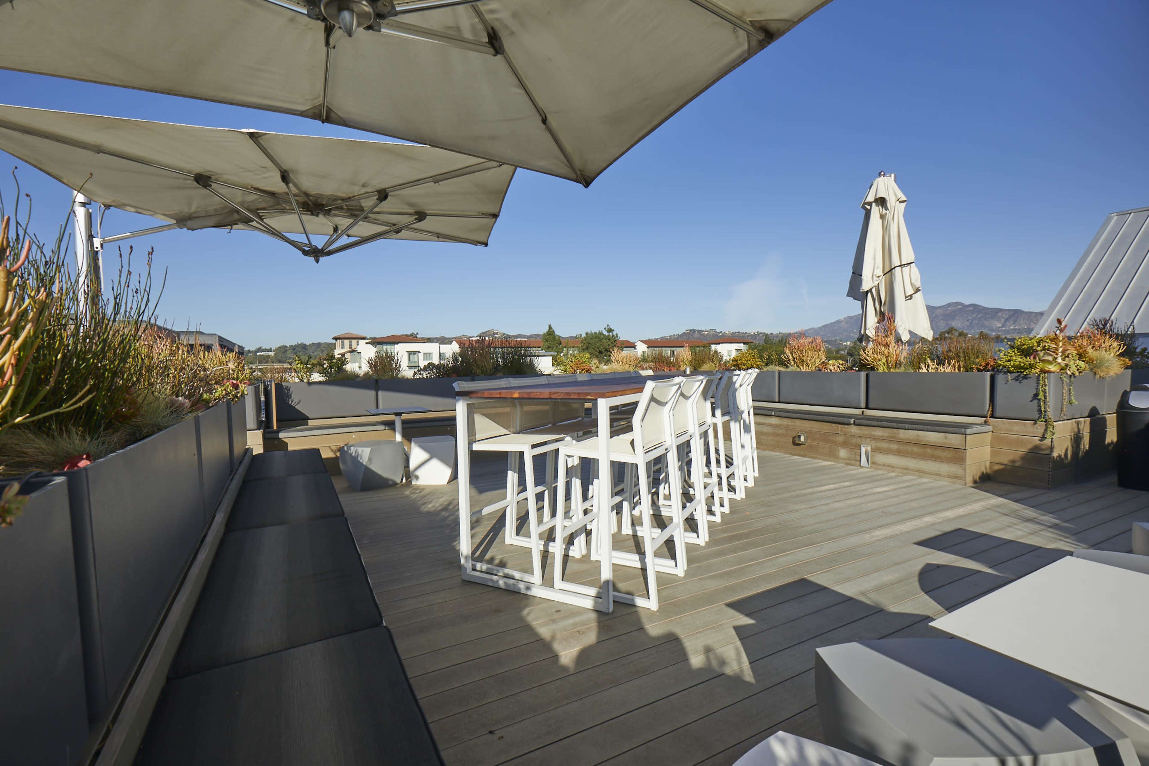 A rooftop patio with seating, large umbrellas, and planters, set against a clear blue sky and distant mountains.