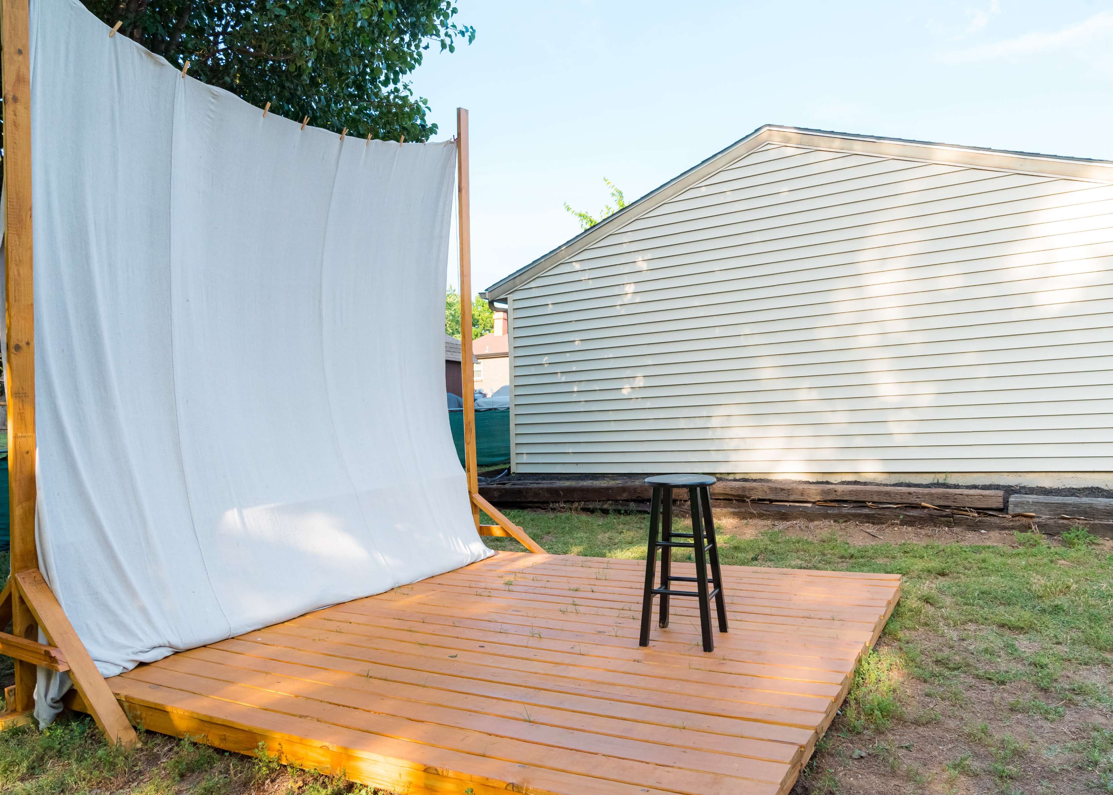A wooden stage with a white backdrop and a black stool stands in a grassy backyard next to a light-colored house.